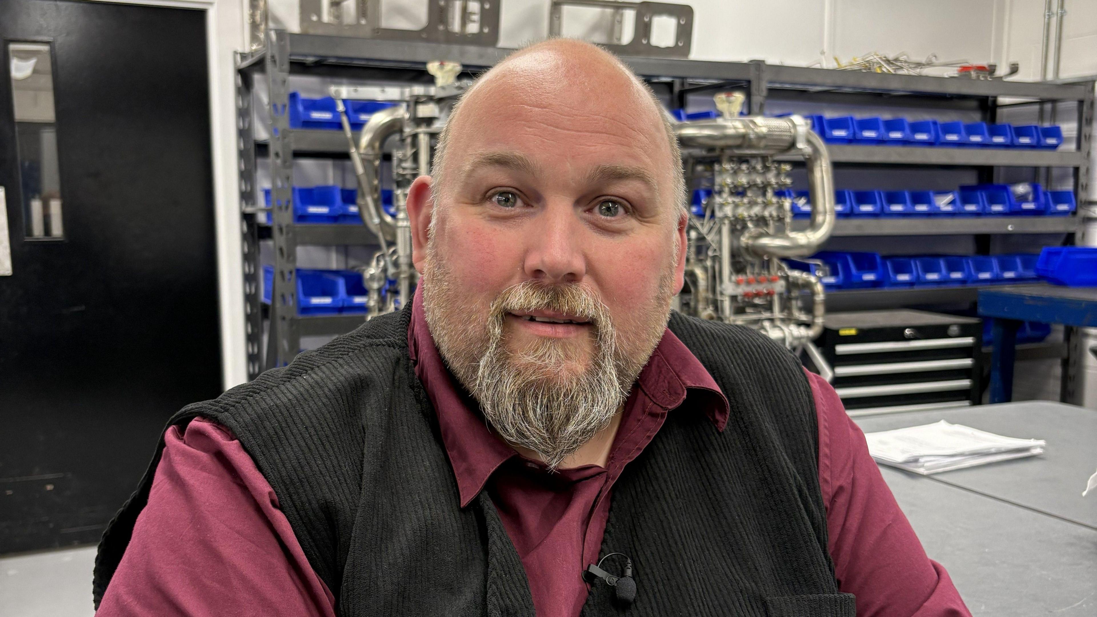 Derek Harris  fom Skyrora, a bald man with a light coloured bears, wearing a black waistcoat and a burgyndy shirt. He is smiling. Behind him are shelves with rows of blue containers and a work table with a metal structure on it.