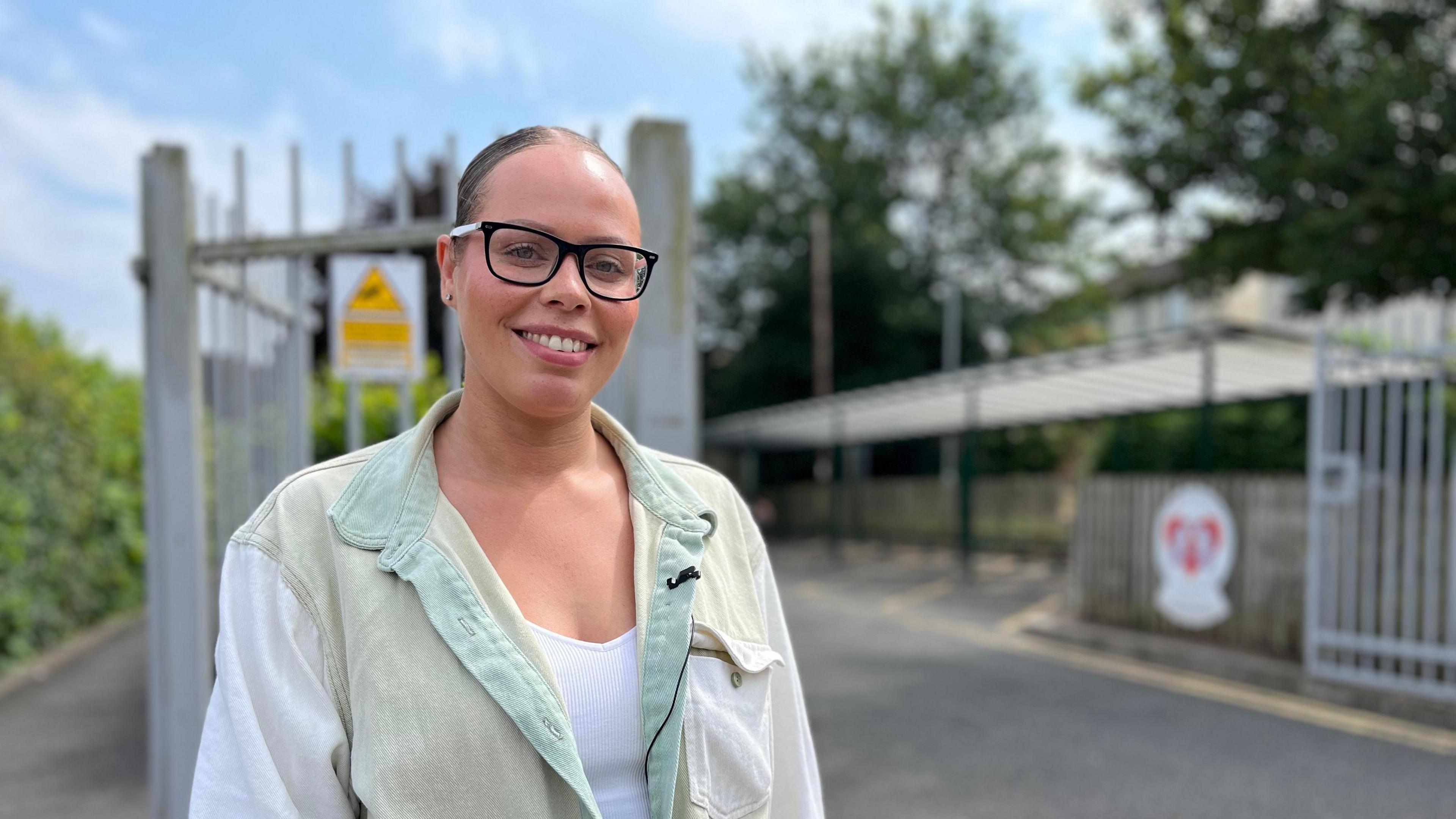 Mother Alicia Bailey stands outside the school gates. She has glasses on and her hair tied back and is wearing a white vest with a green oversized shirt on top.