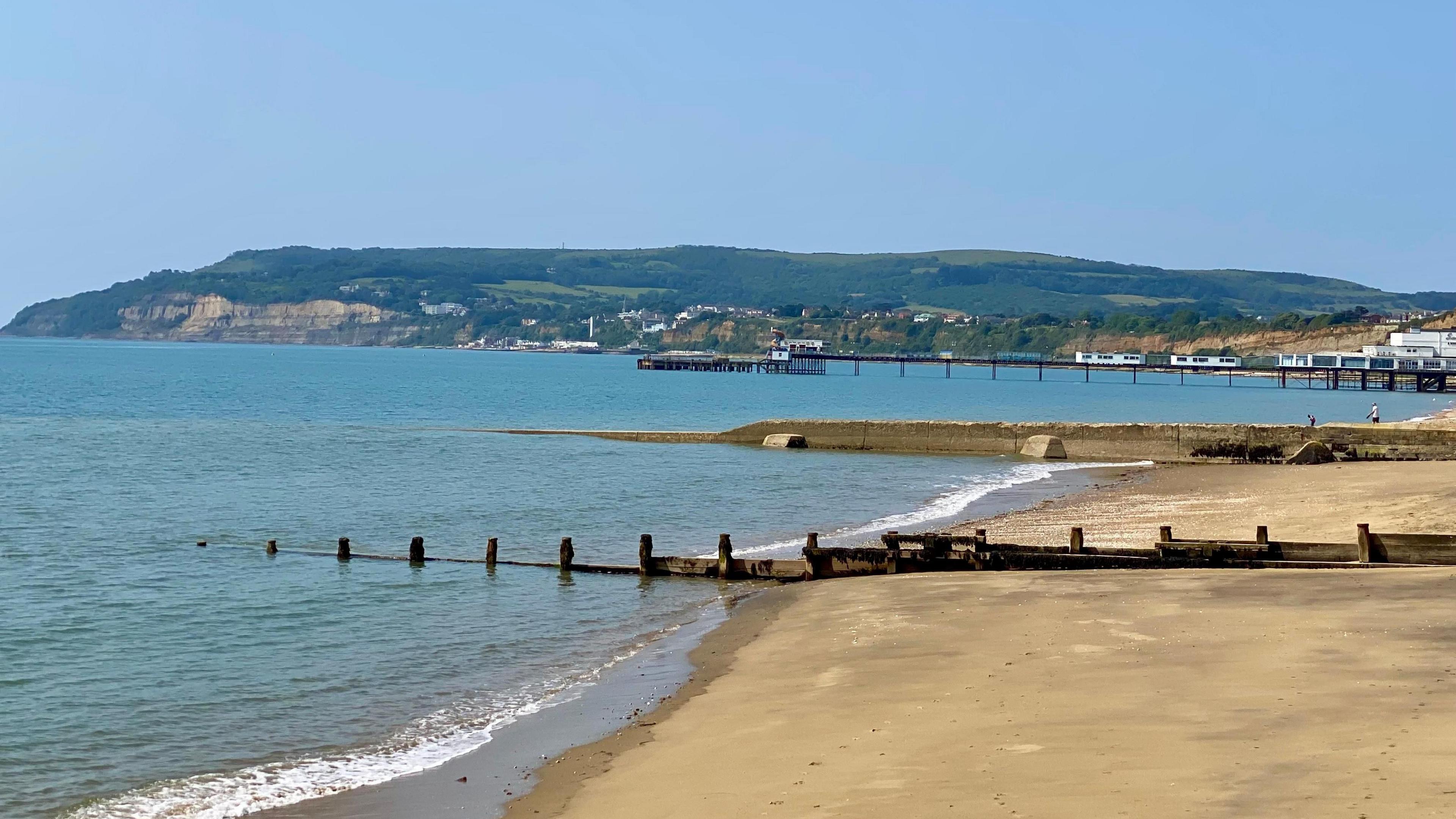 A empty sandy beach and sea on a sunny day with cliffs and green hills in the background.