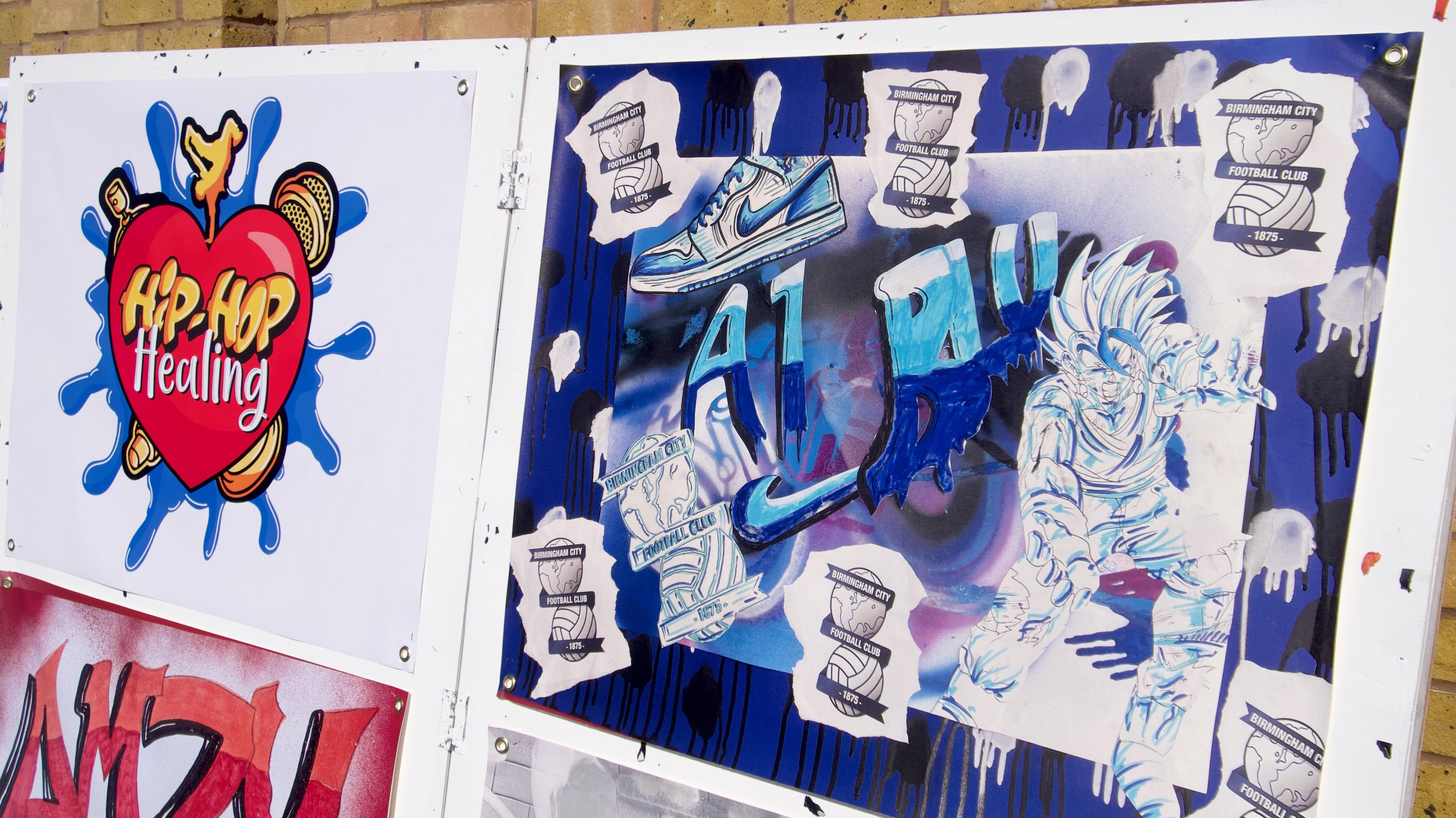 Artwork on a board. One painting has the words hip hop healing on a red heart. The second piece is blue and white and includes six Birmingham City club crests - a globe sat on top of a football