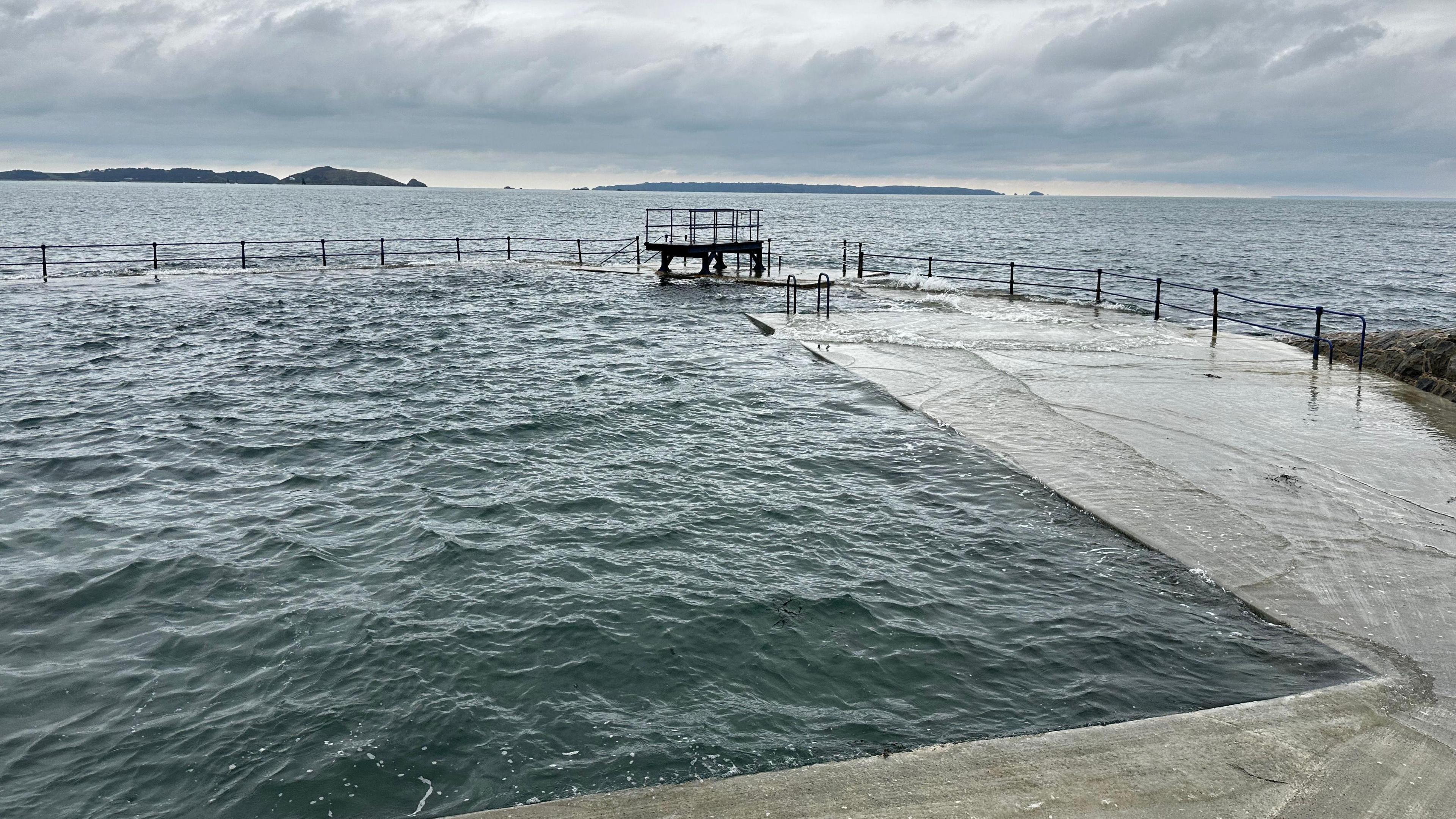 A diving board on the edge of an open air sea water bathing pool on a grey-weather day. Both the sea and pool are at the level of the edge of the pool, and there are islands in the background.