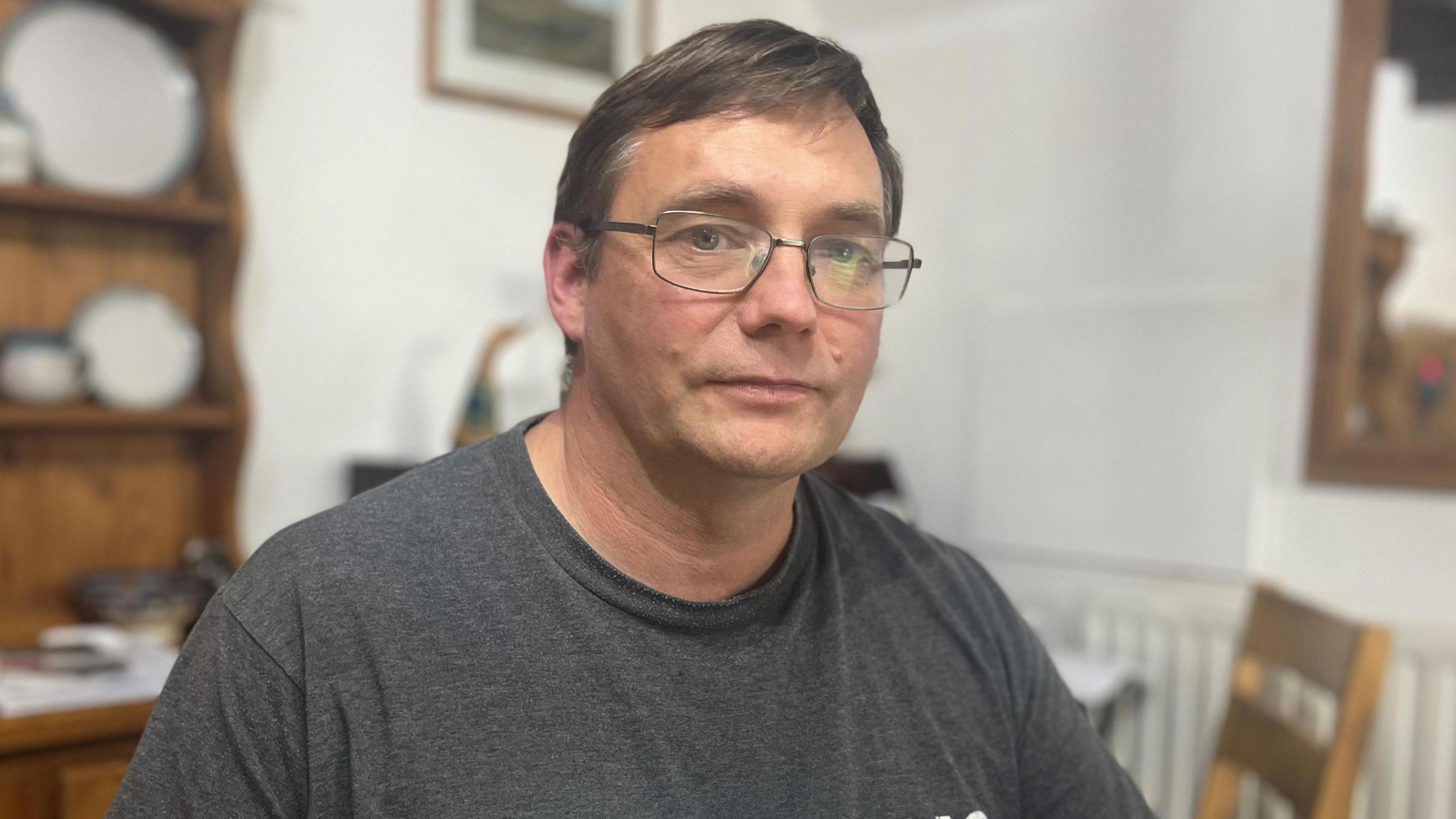 A man wearing glasses and a grey t-shirt sitting in his kitchen