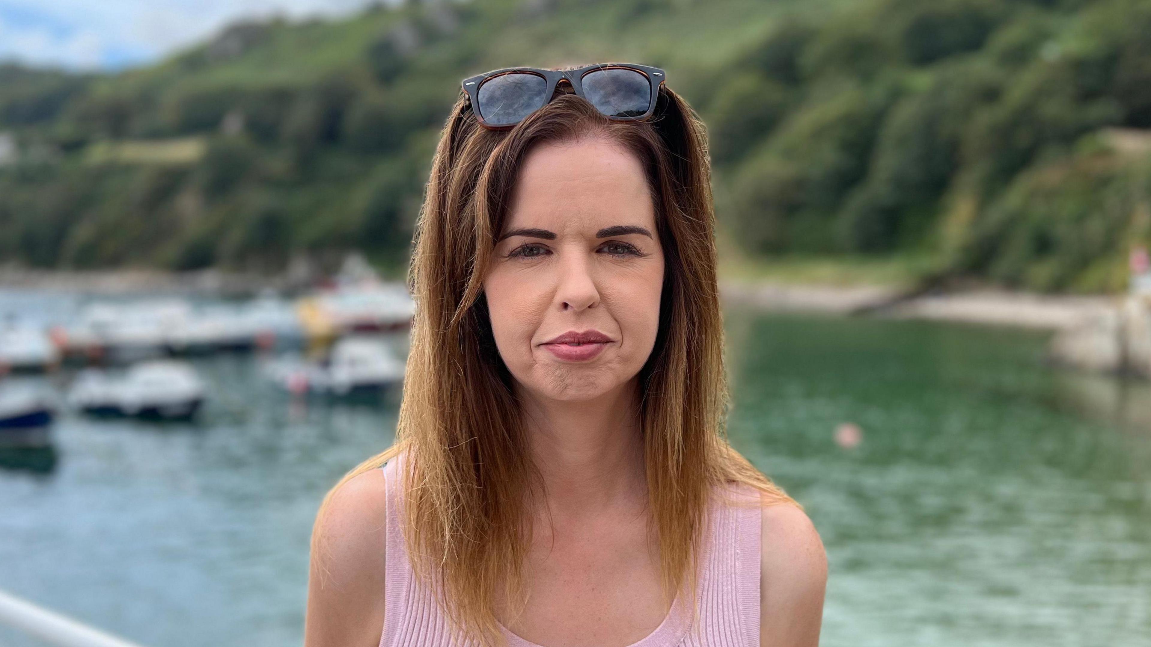 Denise Heavey is standing in front of Bonne Nuit harbour, it is out of focus behind her. She is looking into the camera, wearing a pink tank top with sunglasses on her head.
