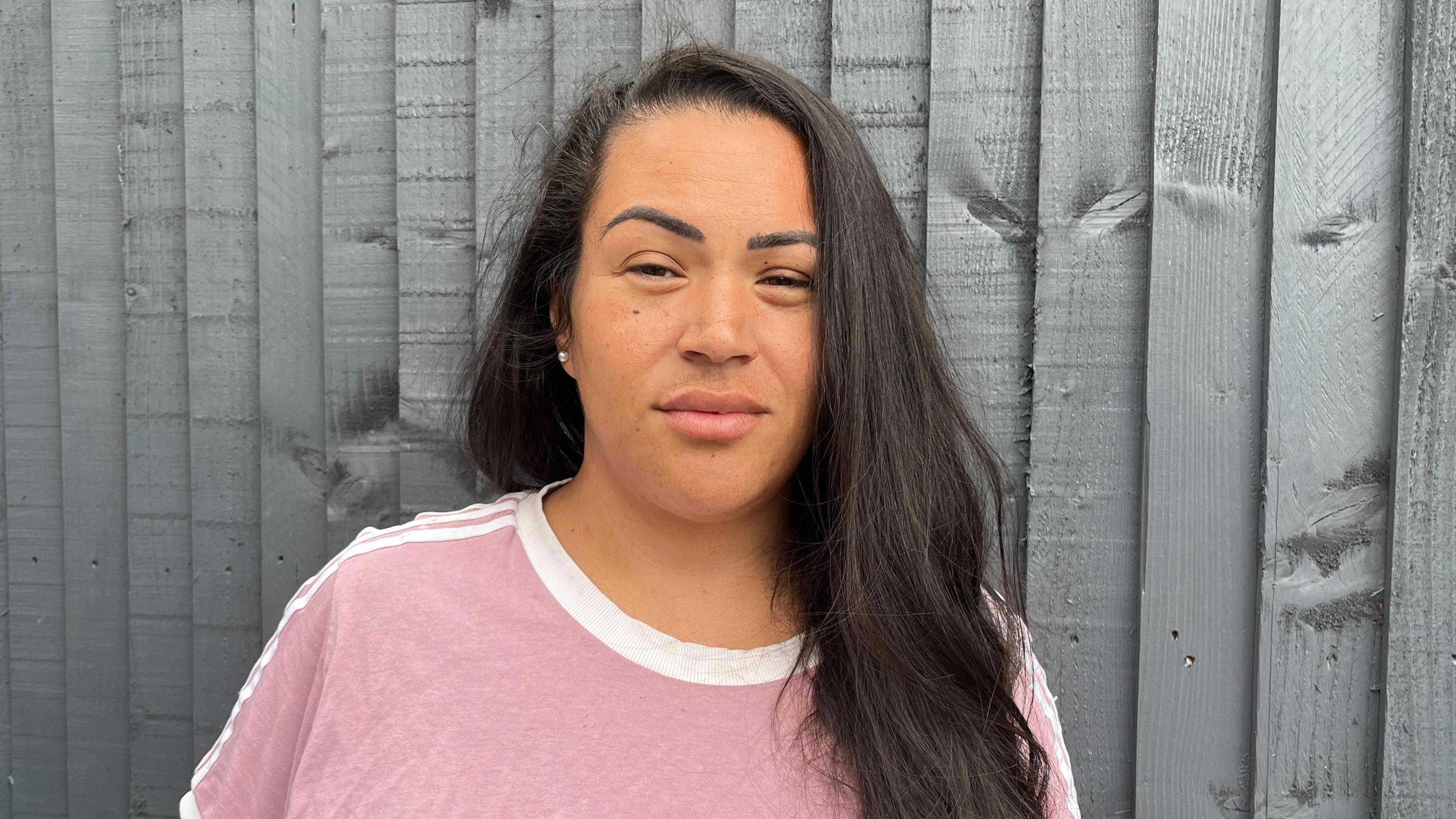 Sasha Field with black long hair looking at the camera. She is in front of a wooden fence and is wearing a pink top.