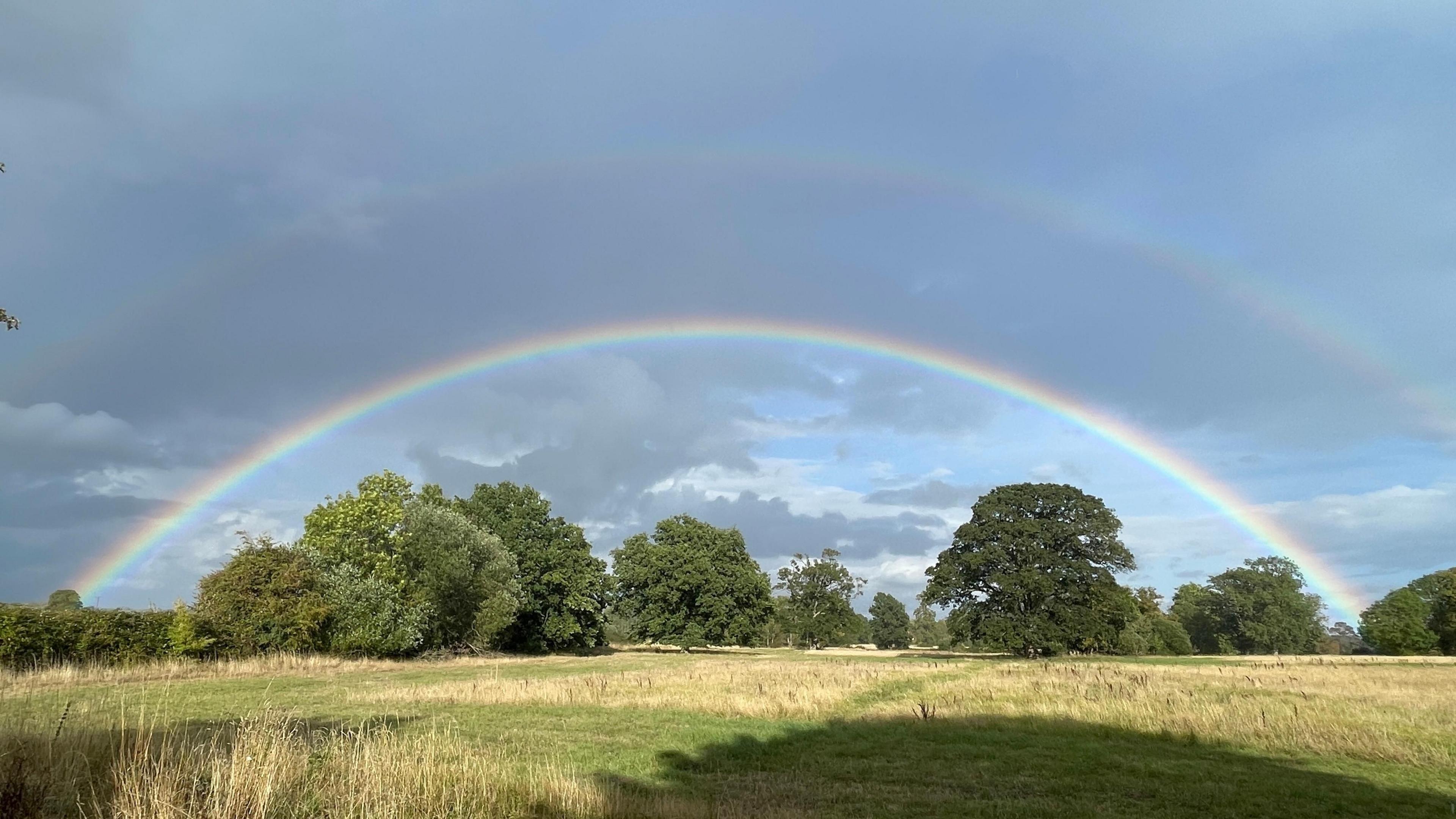 A double rainbow spans the sky above trees. The sun appears to be shining but there are dark clouds in the sky.