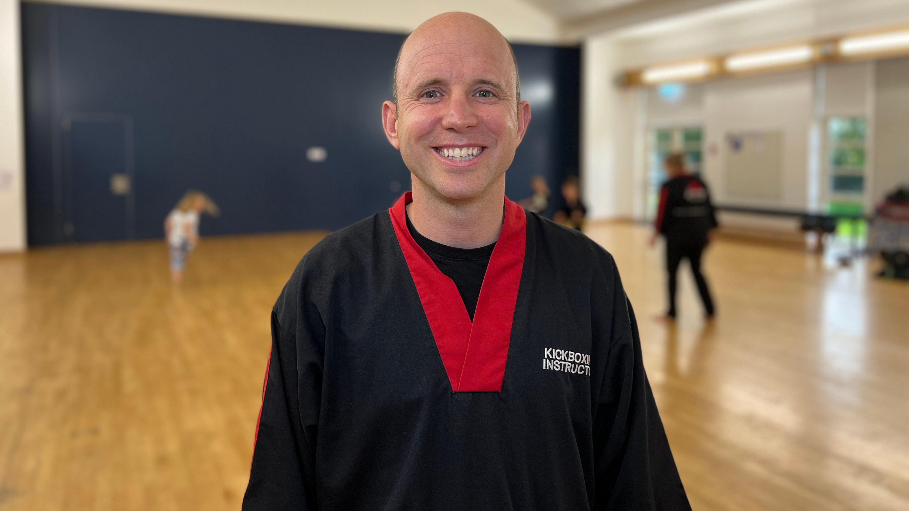 Matt smiles at the camera as in the background a child runs around while another kickboxing coach teaches the class. He is wearing a black karate gi with a red collar and has the words Kickboxing Instructor on his chest. Matt is wearing a black t-shirt underneath his gi and he is bald.