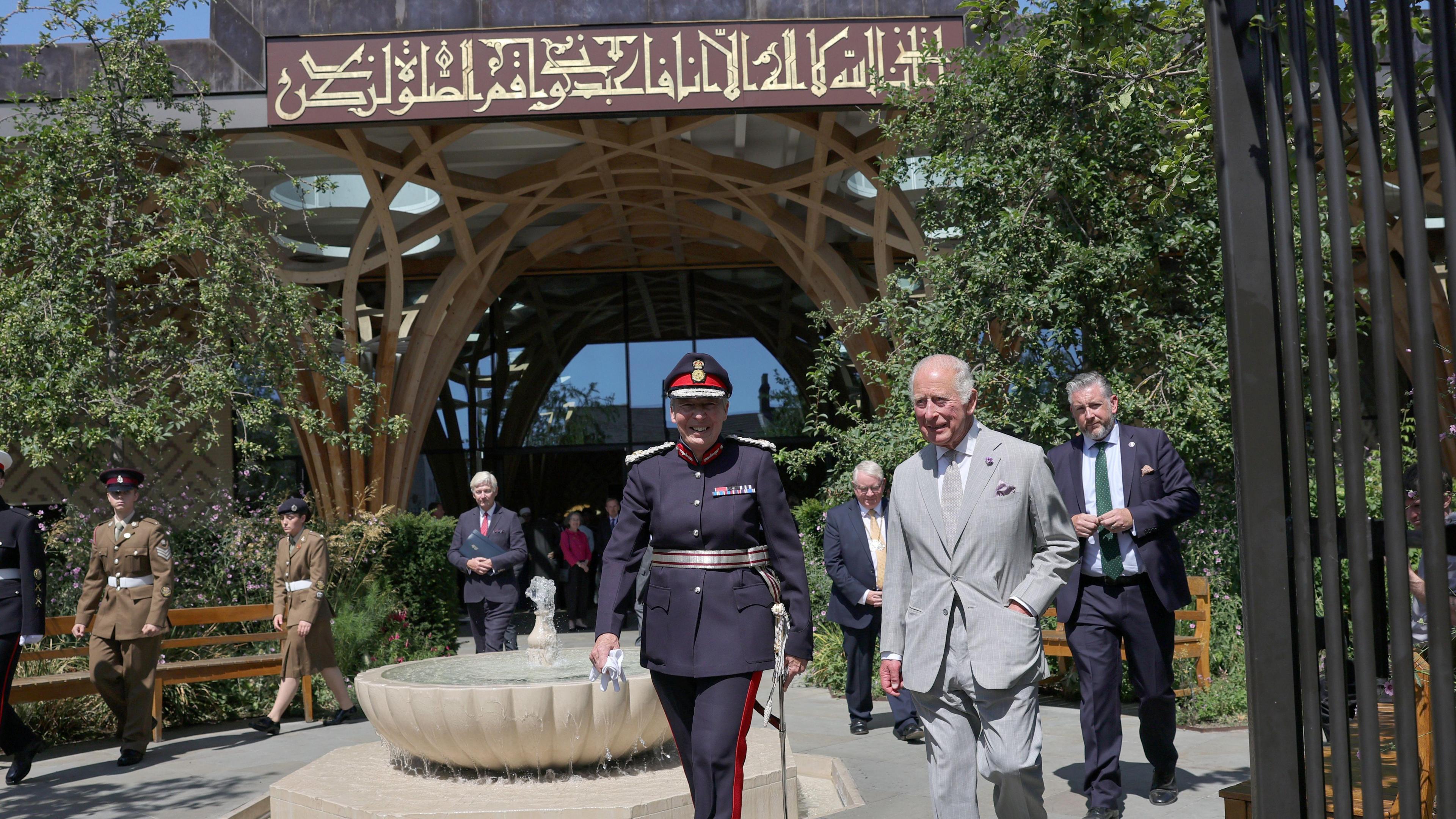 King Charles III and someone in military uniform are walking towards a gate which is in front of a large mosque which has scripture above its entrance.