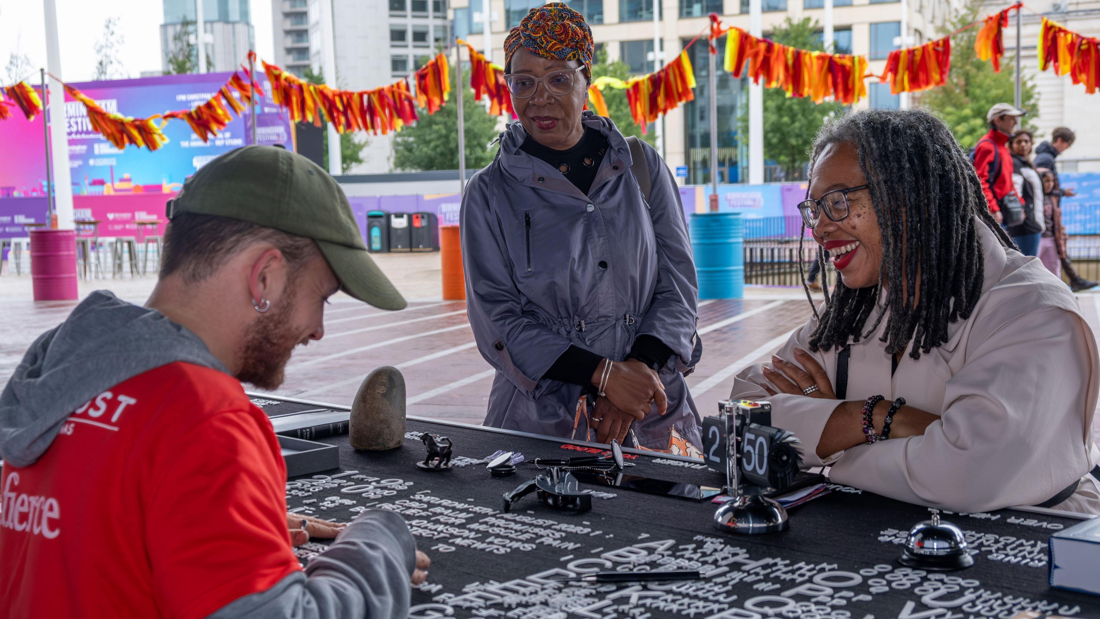 A man, wearing a red and grey top, and a green hat, smiling to two women, who are taking part in a cultural art event