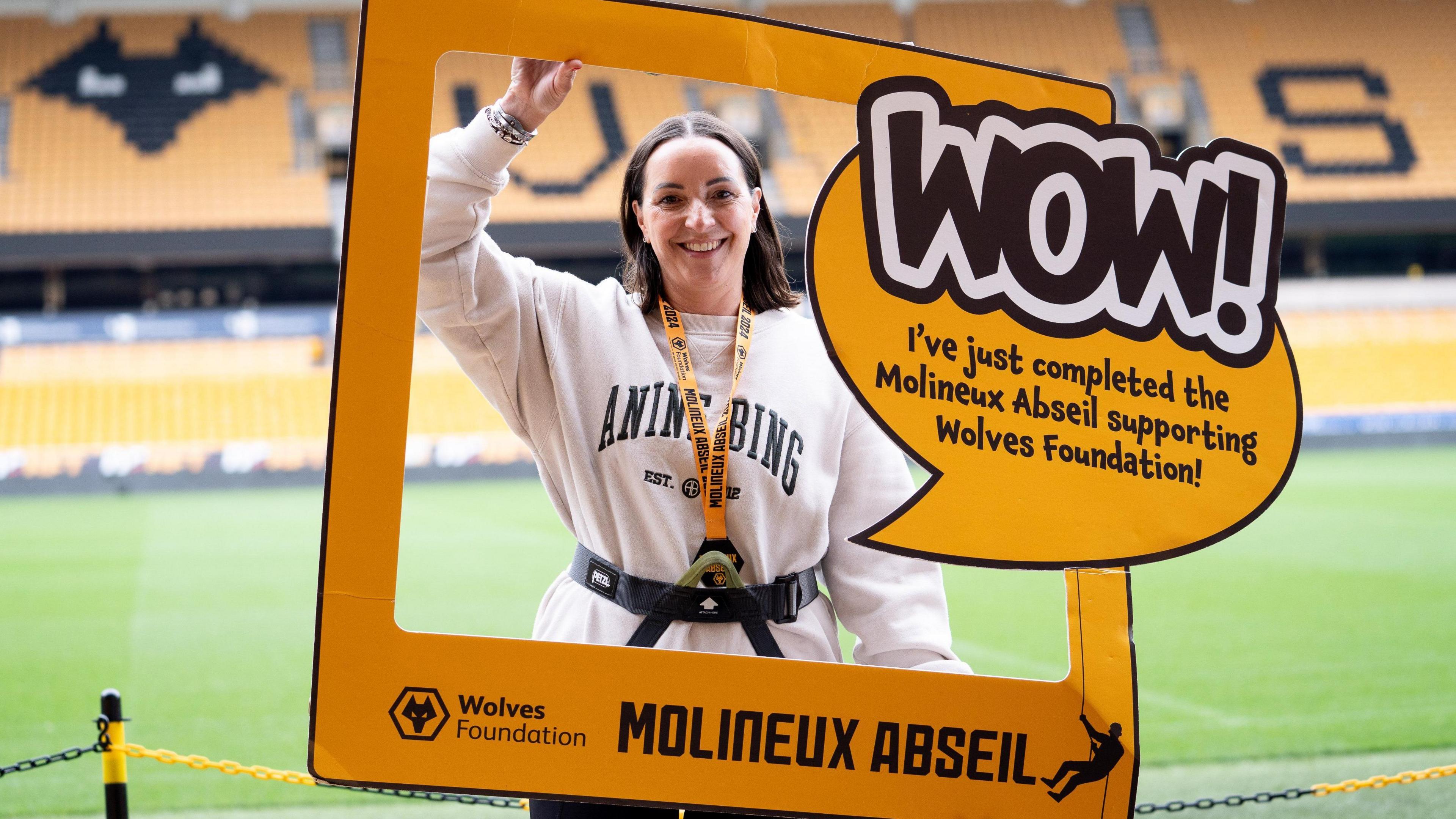 A woman with brown hair and a white jumper holding an orange picture frame that says 'Wow! I've just completed the Molineux Abseil supporting Wolves Foundation!" She is standing in the stadium grounds. 