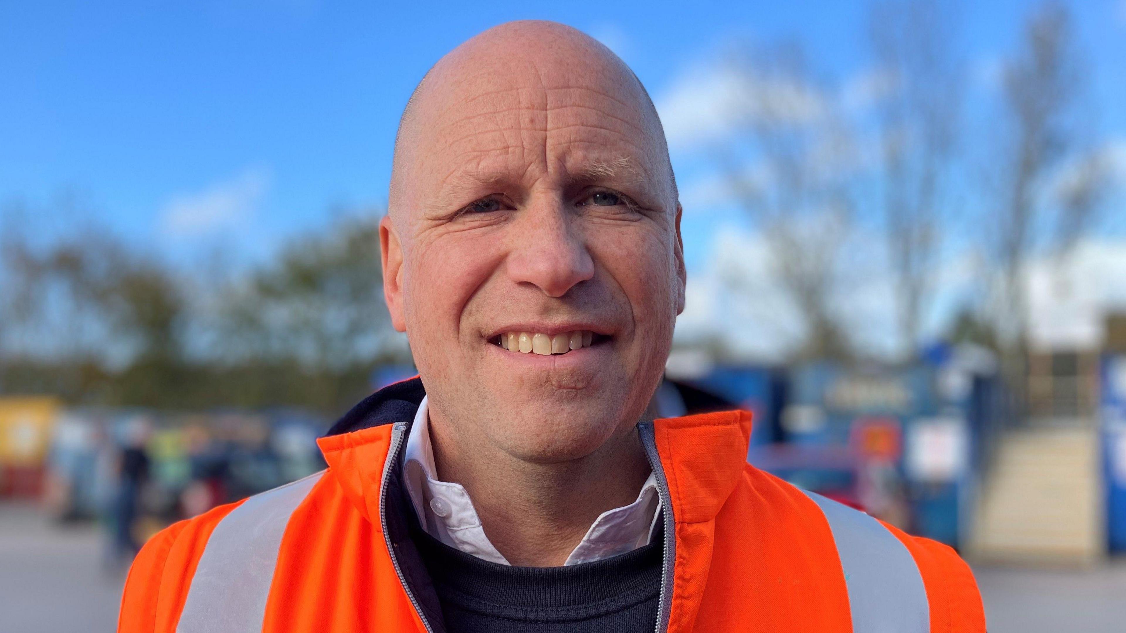 Bald man wearing a high viz jacket and trying to smile at the camera looking very unsure