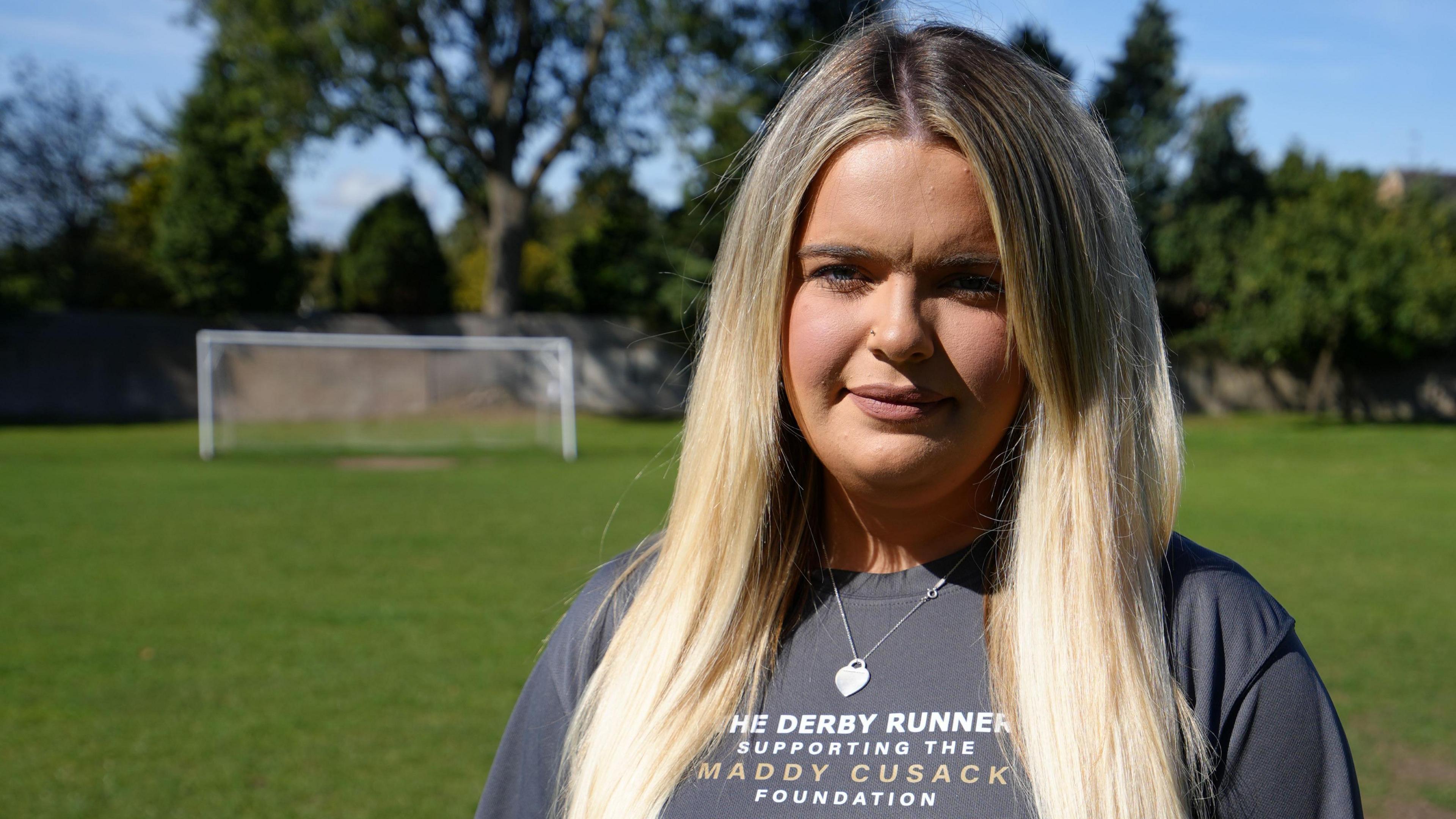 Olivia Cusack pictured at a football field in Derby on a sunny day. Olivia has long blonde hair worn loose and wears a grey T-shirt promoting the Maddy Cusack Foundation. Behind her is a line of trees and a goal. 