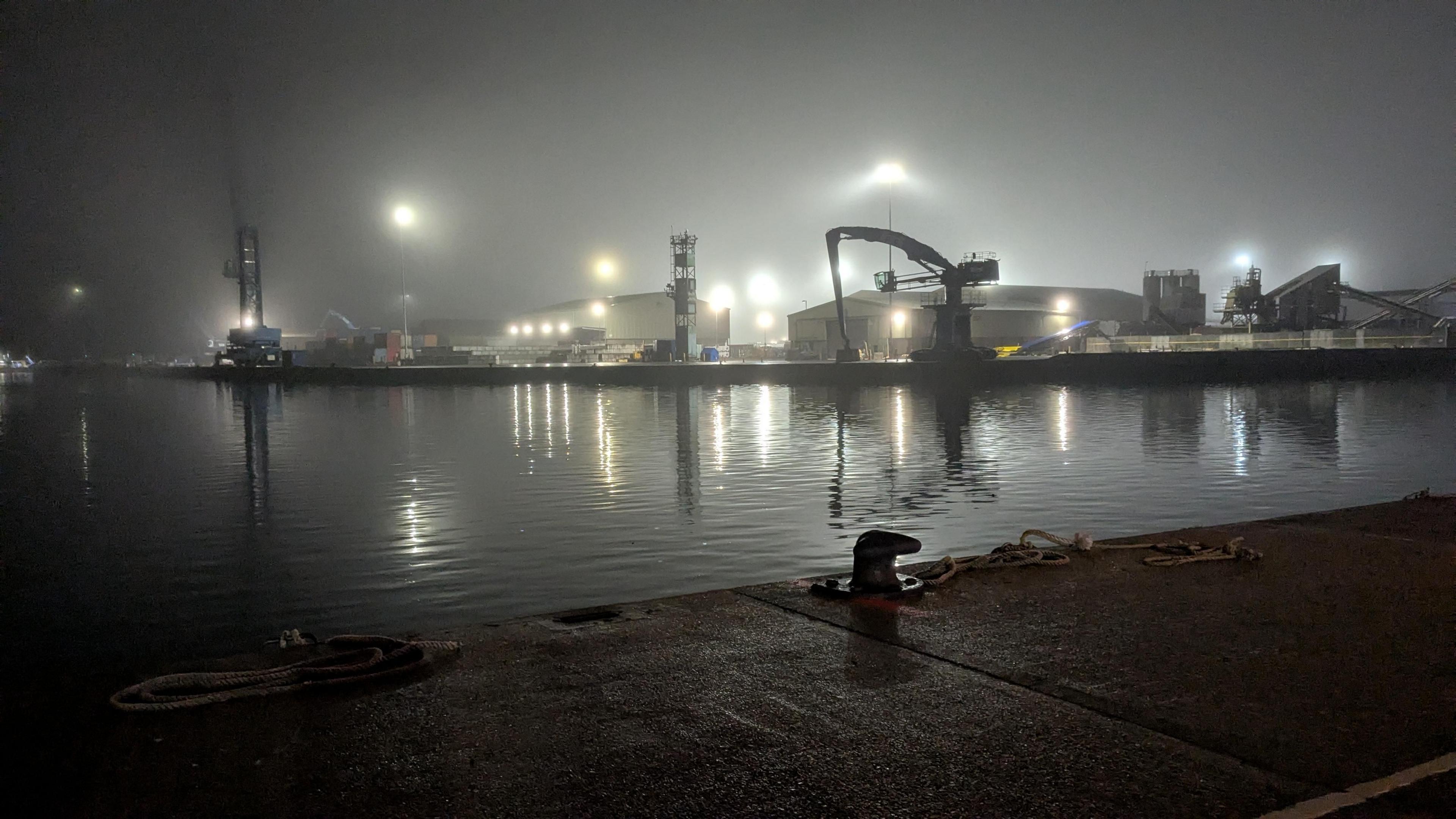 Poole quay at night in the mist. There are large floodlights over cranes and the light is reflected in the water of the harbour. 