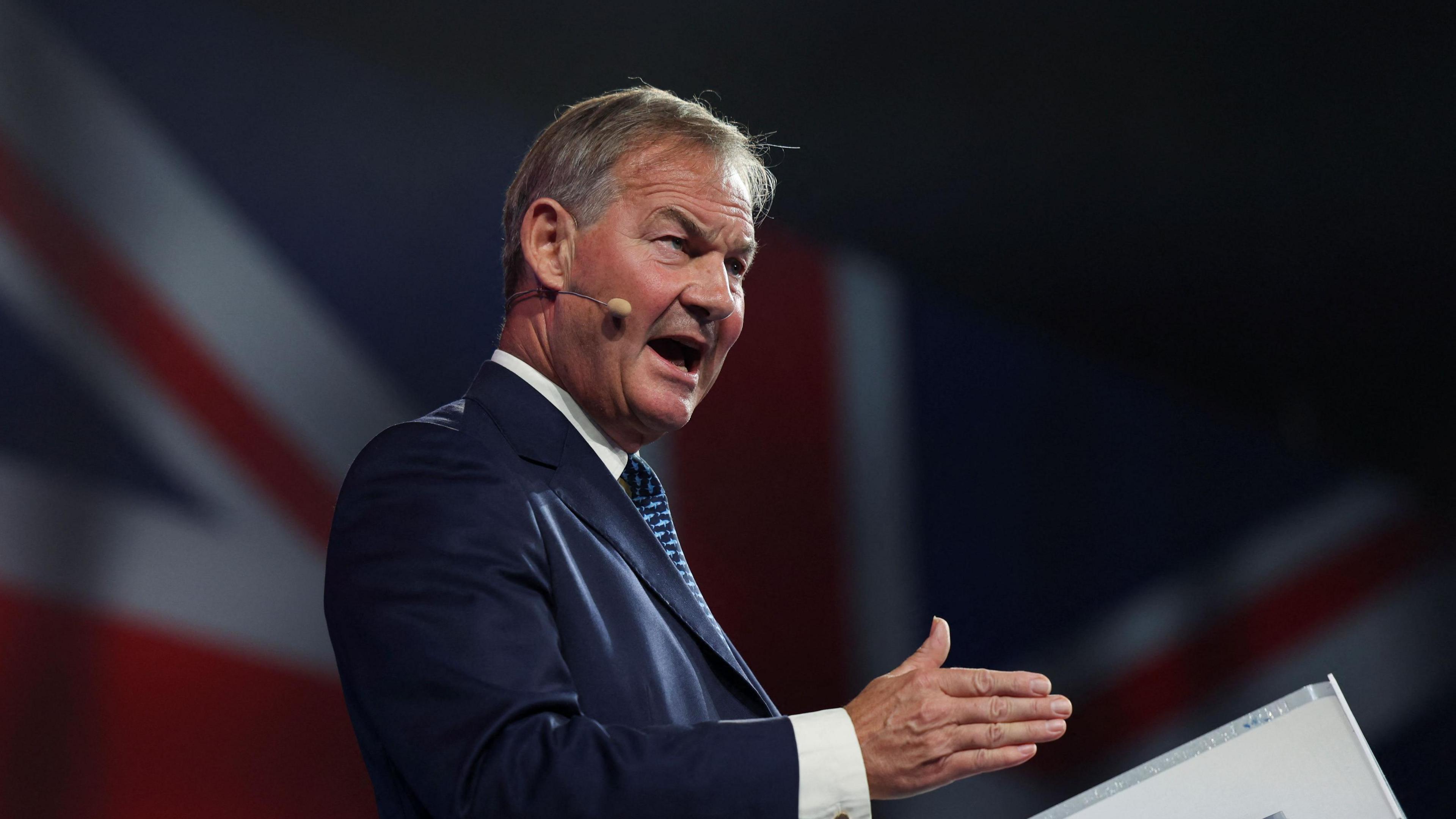 Rupert Lowe is standing at a podium giving a speech. He's wearing a dark blue jacket, white shirt and blue striped tie. Behind him, we can see a Union Flag.