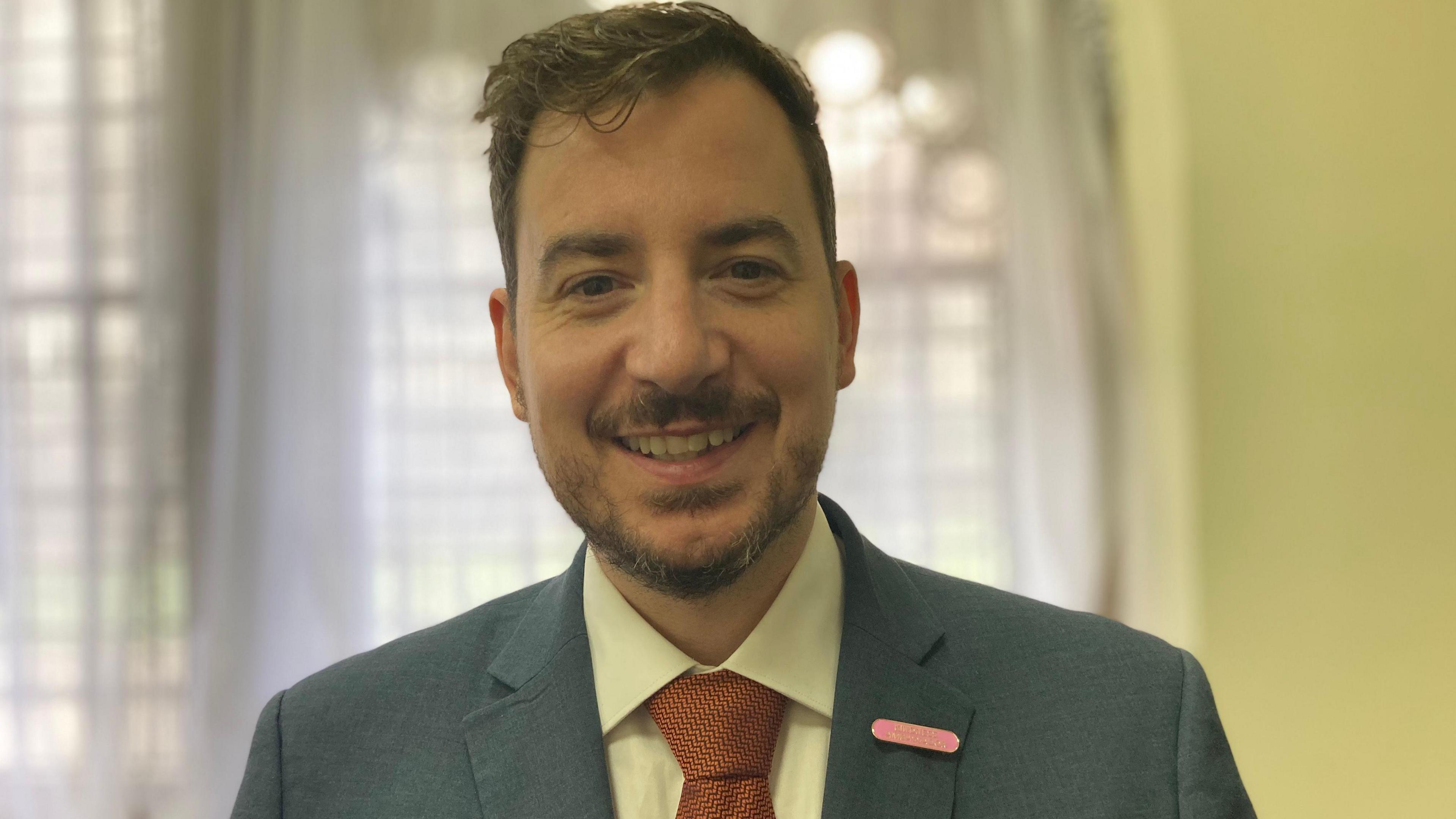 Mike Reader in a light blue suit and a white shirt with a red tie smiles in a head-and-shoulders shot.