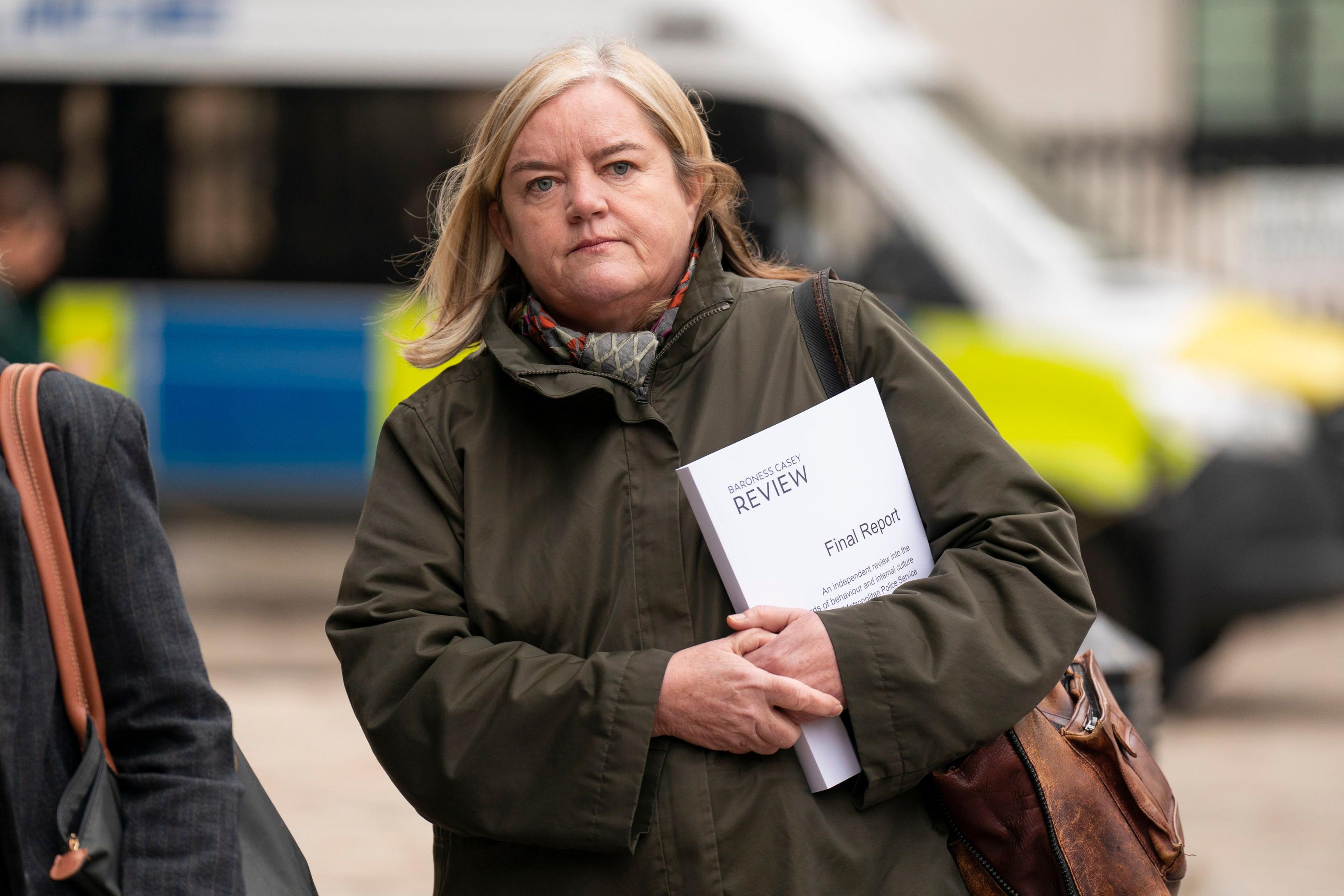 Baroness Casey clutches a copy of her report into the behaviour and culture at the Metropolitan Police Service. She is walking towards the camera wearing a green, waxed jacket and is carrying a leather shoulder bag.
