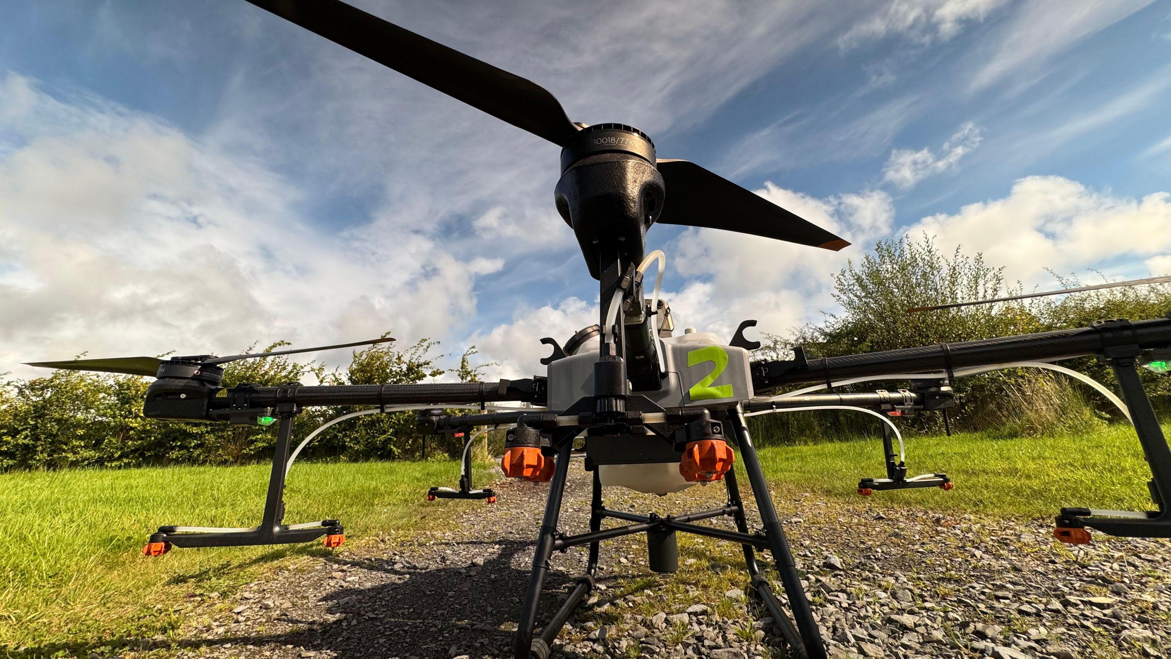 Drone on the ground outside in the sunshine. It has four black arms with orange stabilisers. The drone is marked with a green 2 on a grey box. The propeller on top is black.