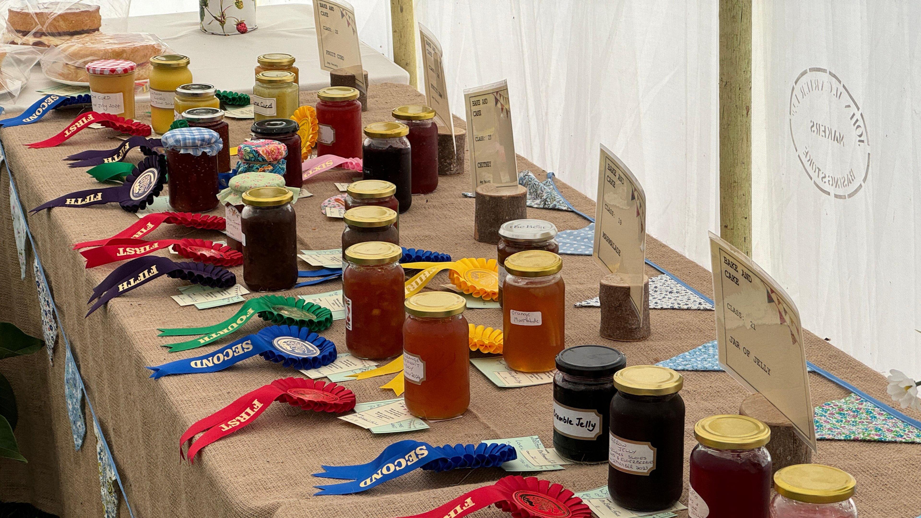 A table with a straw table cloth and jars of jelly marmalade and jam on it. There are different rosettes in front of each jar.