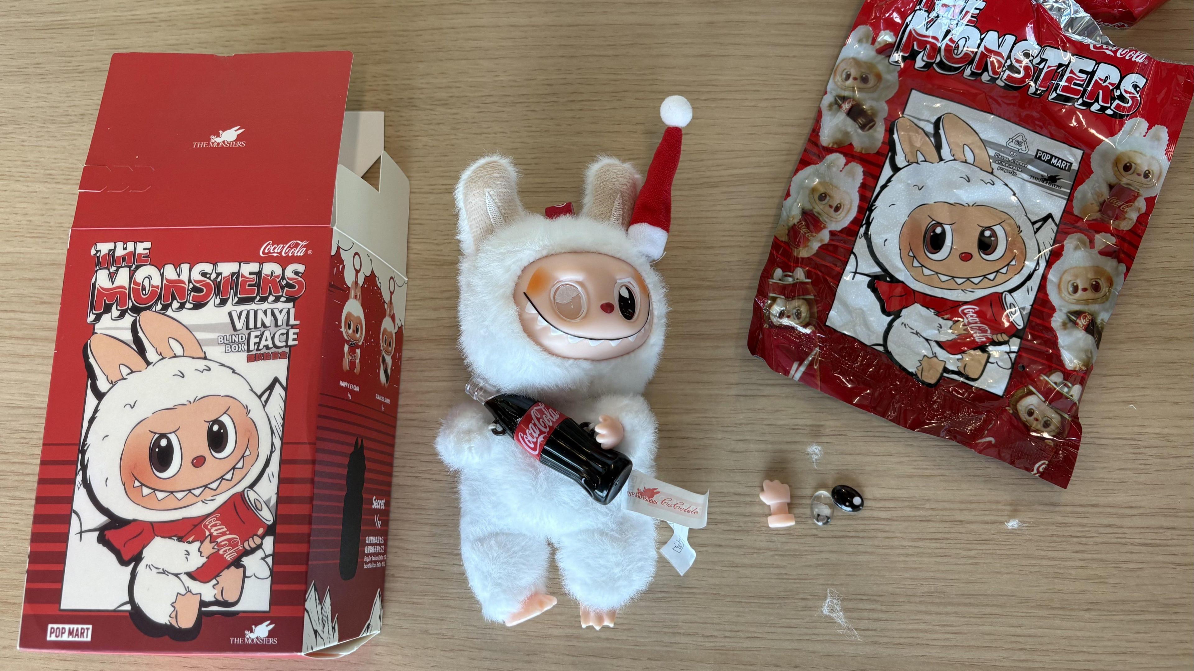 A fake Labubu doll rests on a wooden surface. It has a white plush body and plastic face. It is missing one eye. It holds a small Coca Cola bottle. One of its hands which has fallen off and its missing eye rest next to it along with a packaging box and plastic packaging bag.