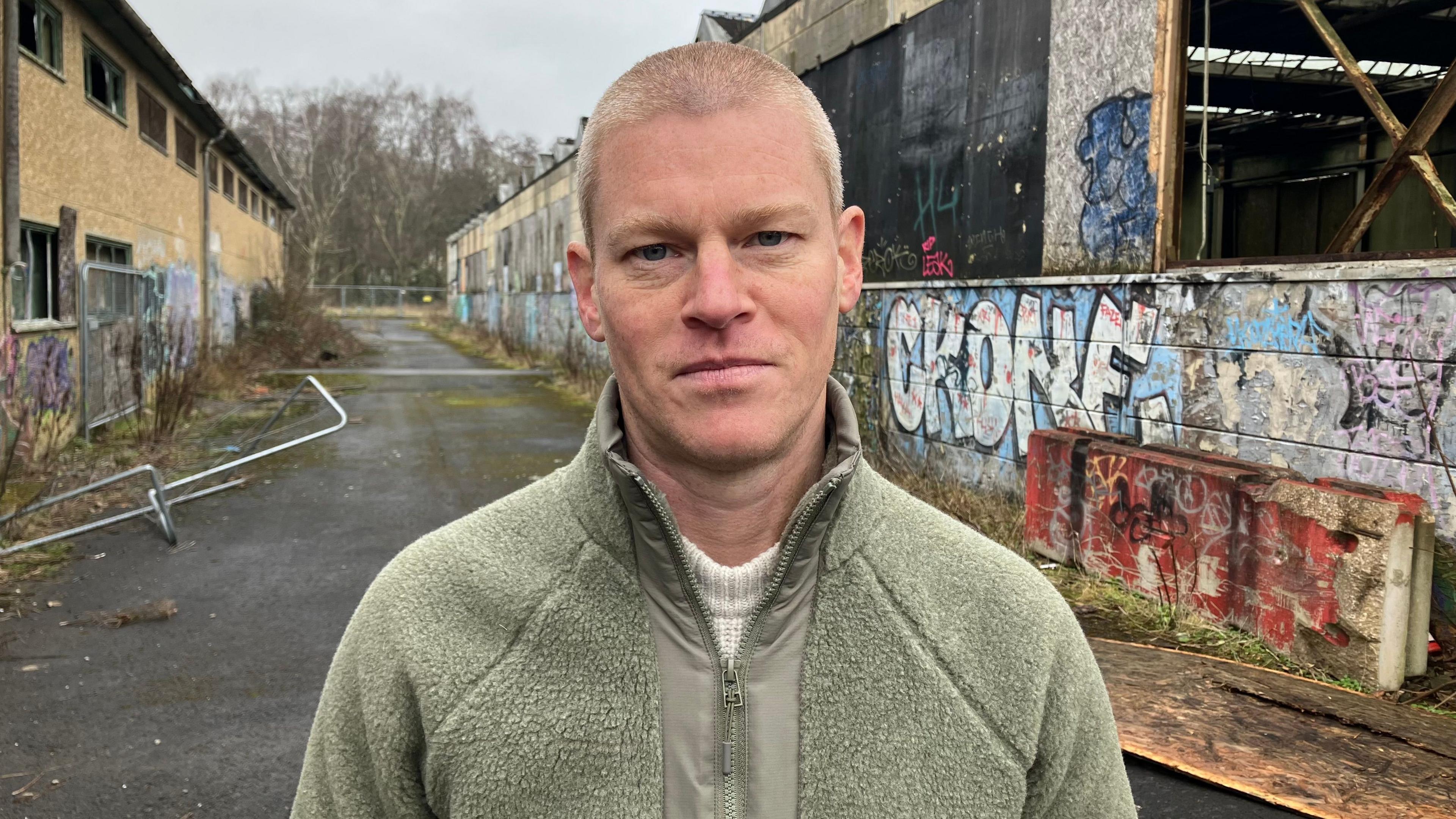 Paul Oster has a short buzz cut with light hair. He is wearing a light green zipped jacket. He is standing. Behind him is the derelict site.