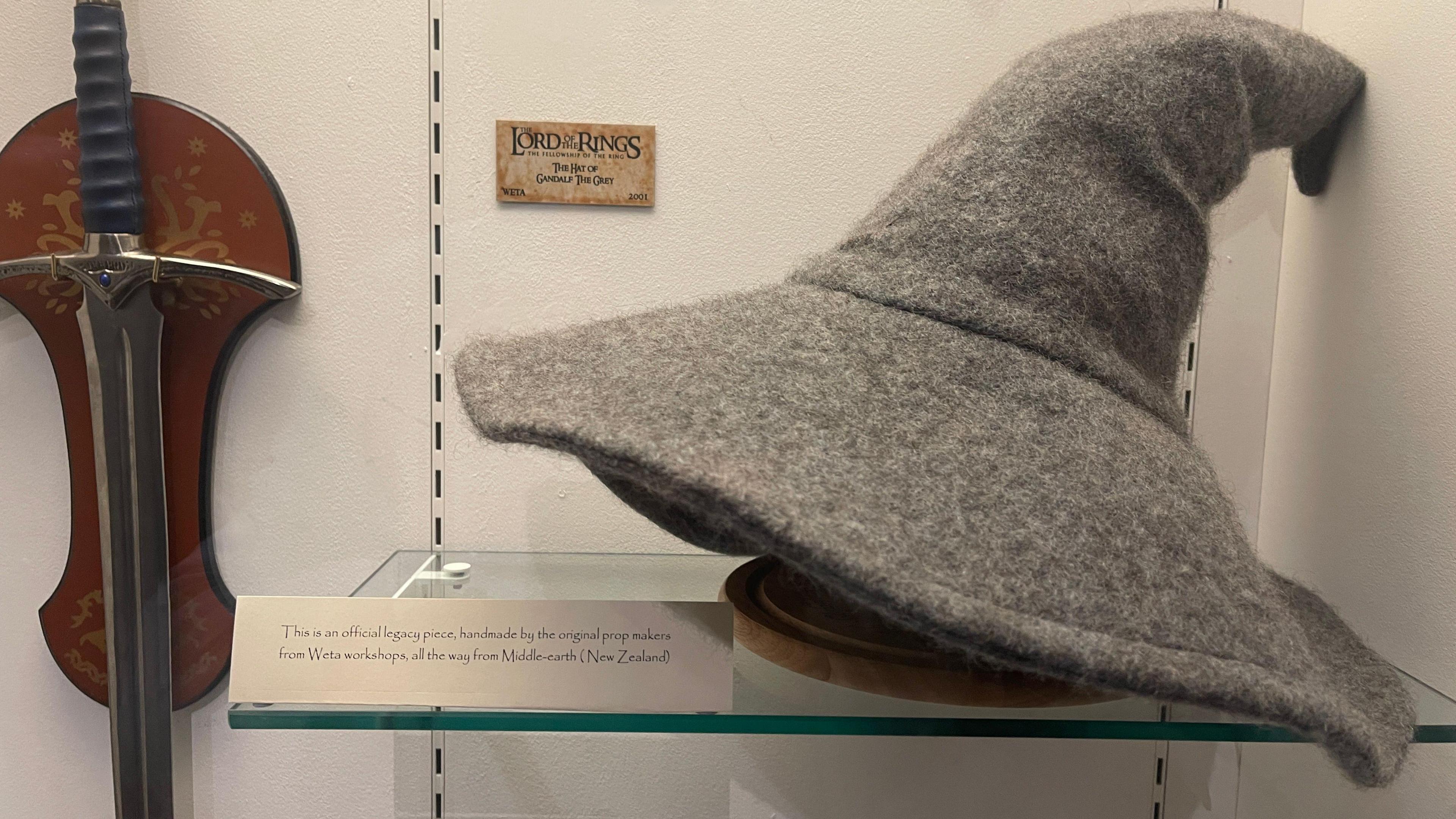 A large grey wizard's hat from Lord of the Rings on a glass shelf next to a small plaque describing what it is and a sword attached to the wall.
