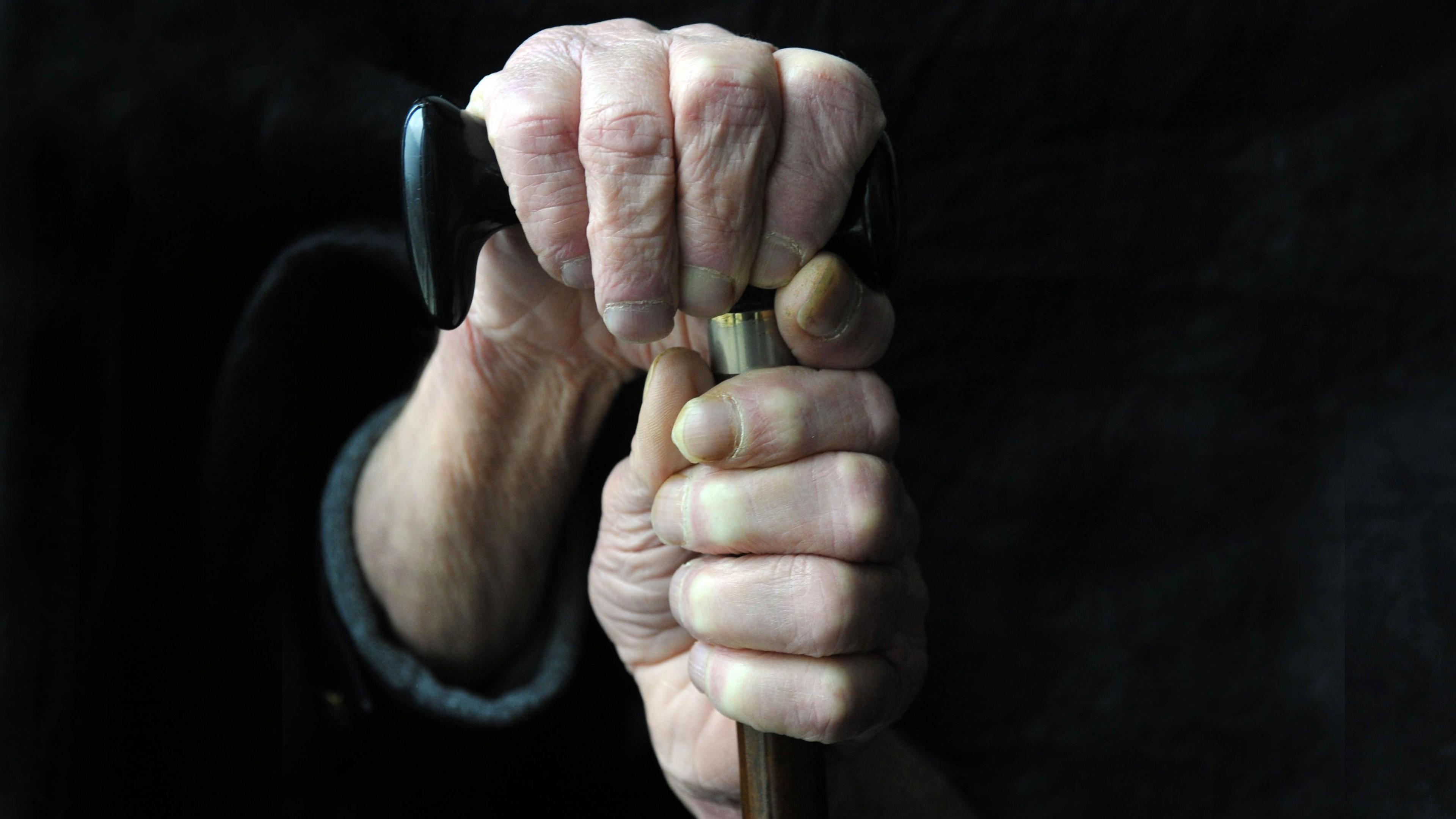 An elderly mans hands is seen grasping the top of his walking stick, with black background. 