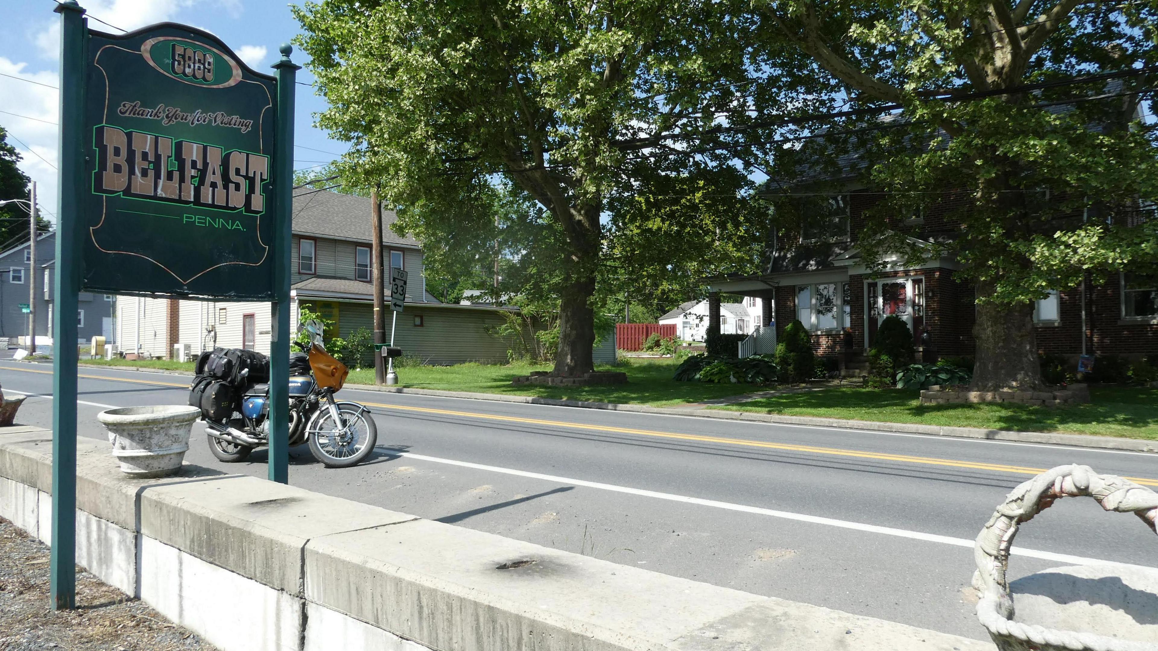 A green sign with Belfast on it in a small town in America, there are trees and three houses beside the road and a motorbike is parked there 