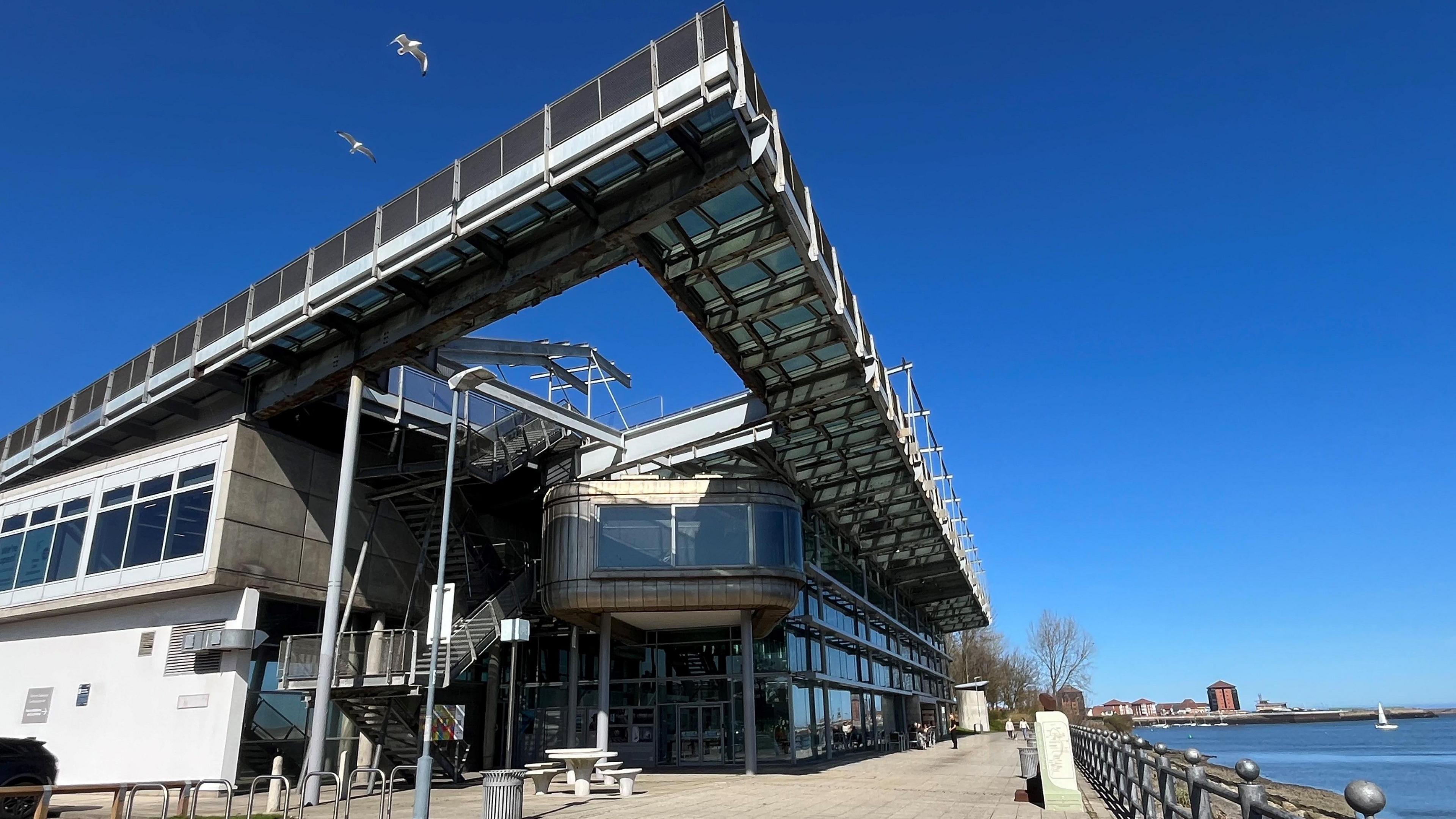 The National Glass Centre is a tall building made of metal panels and glass, overlooking the River Wear. There is seating space and a cafe in front of it, with a dog walker in the distance.