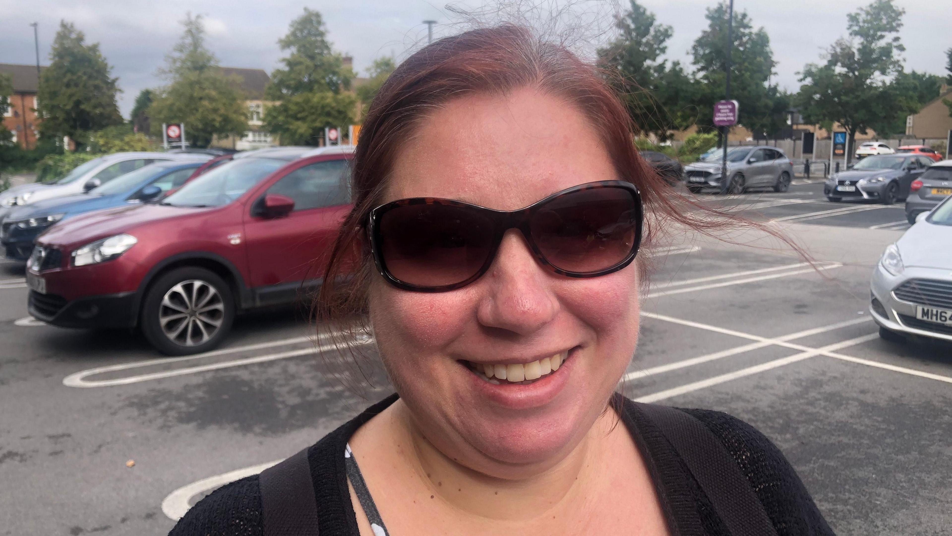 Clare Court, is standing in a car park wearing sunglasses and a black top. 
She has brown hair and is smiling at the camera.