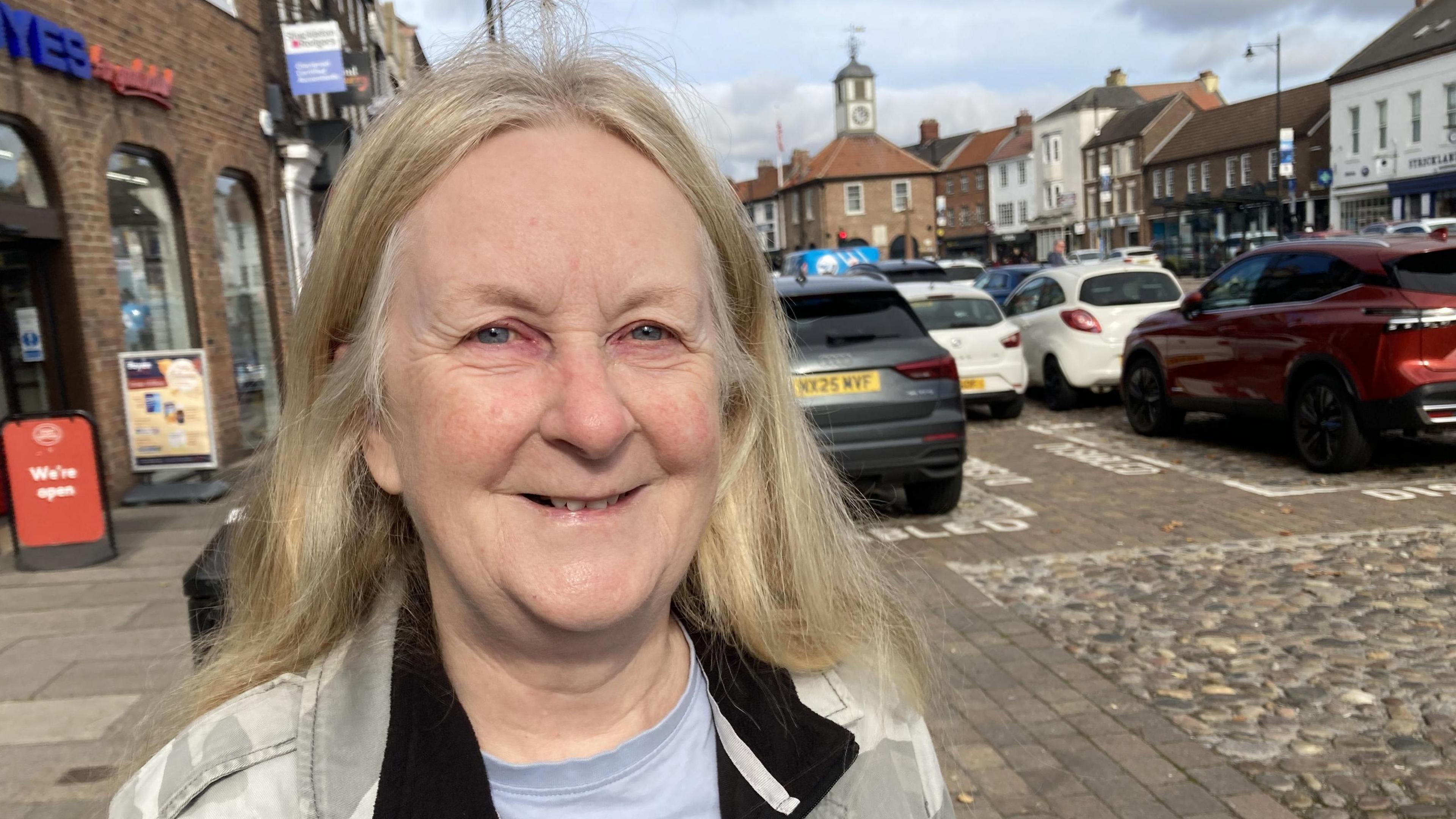 Carole Jones, who has blonde hair, wearing a blue top and grey camouflage coat, looks at the camera as she stands on a pavement on a high street. Parked cars are on the cobbled road next to her.