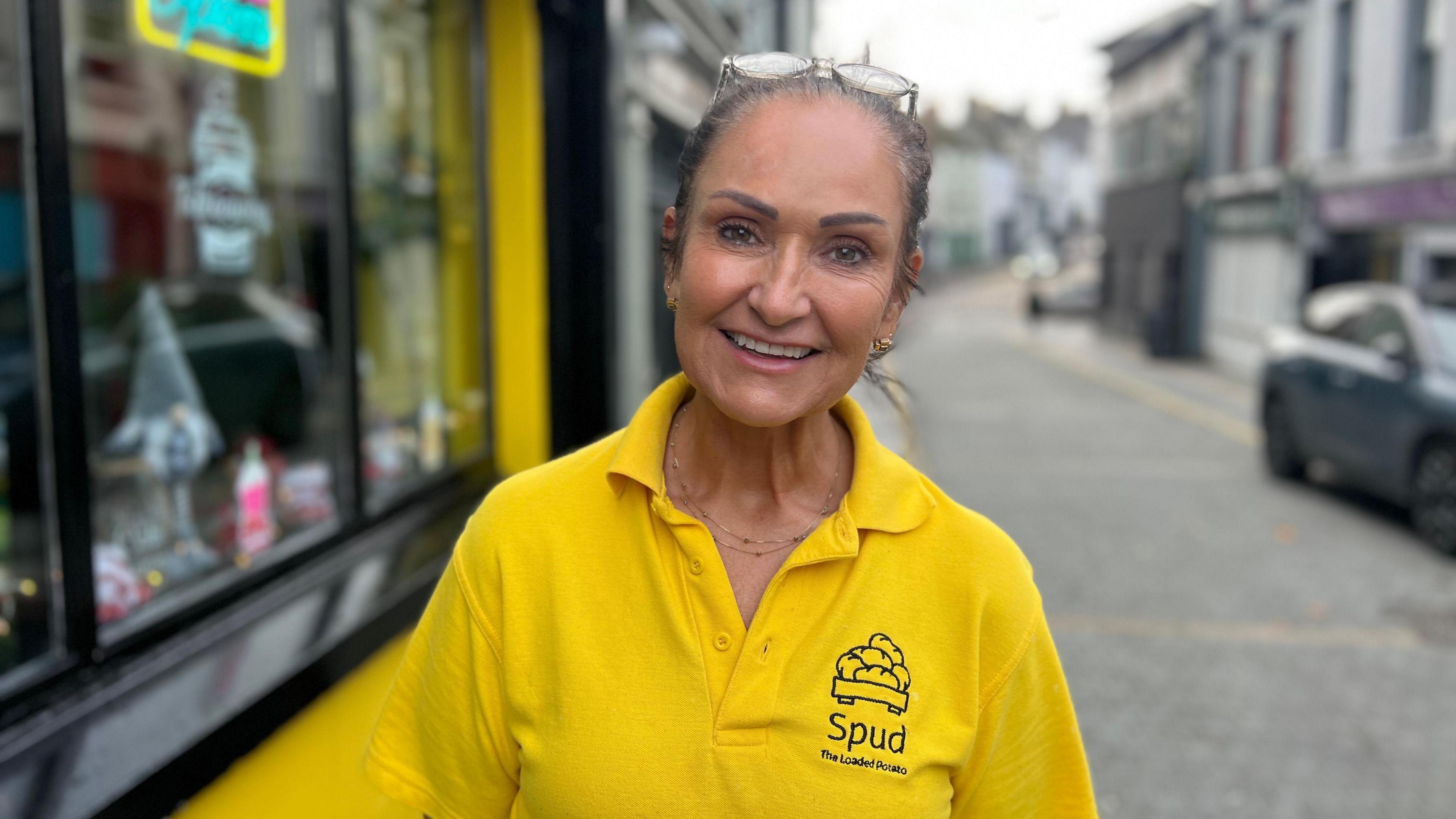 Helen Martin is wearing a yellow t-shirt. Glasses are perched on top of her head. Her brown hair is tied back. Behind her, is a cafe.