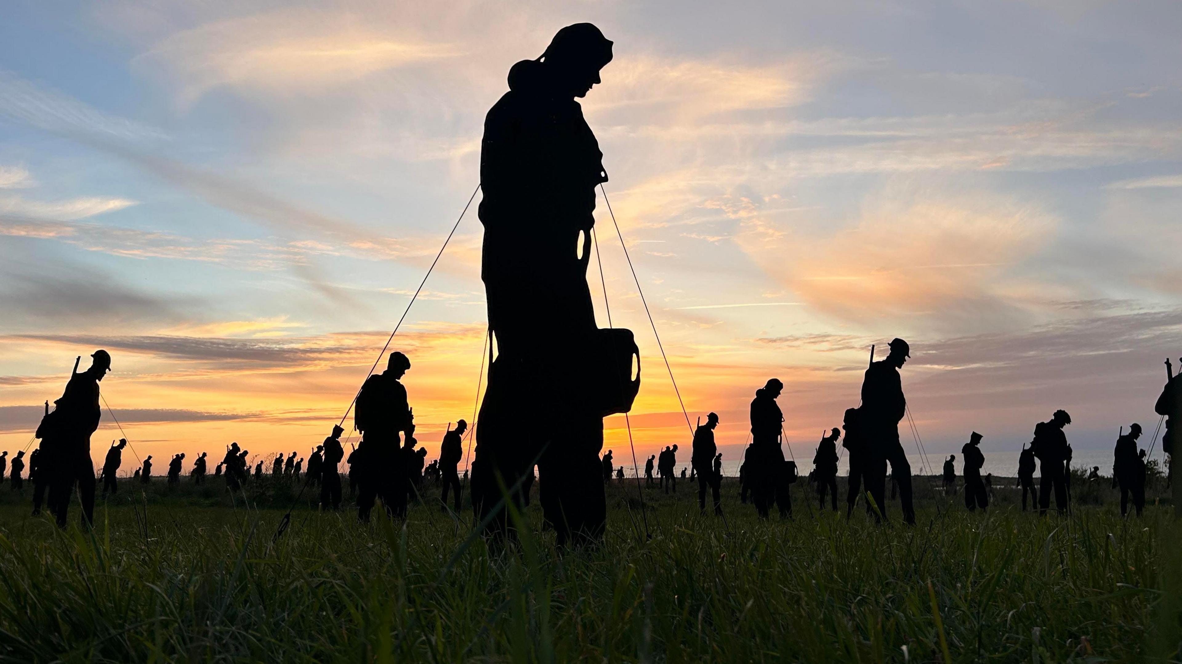 A large number of black silhouettes of servicemen in a garden. You can see grass in the front and a sunset behind. An airman figure is in the foreground. 