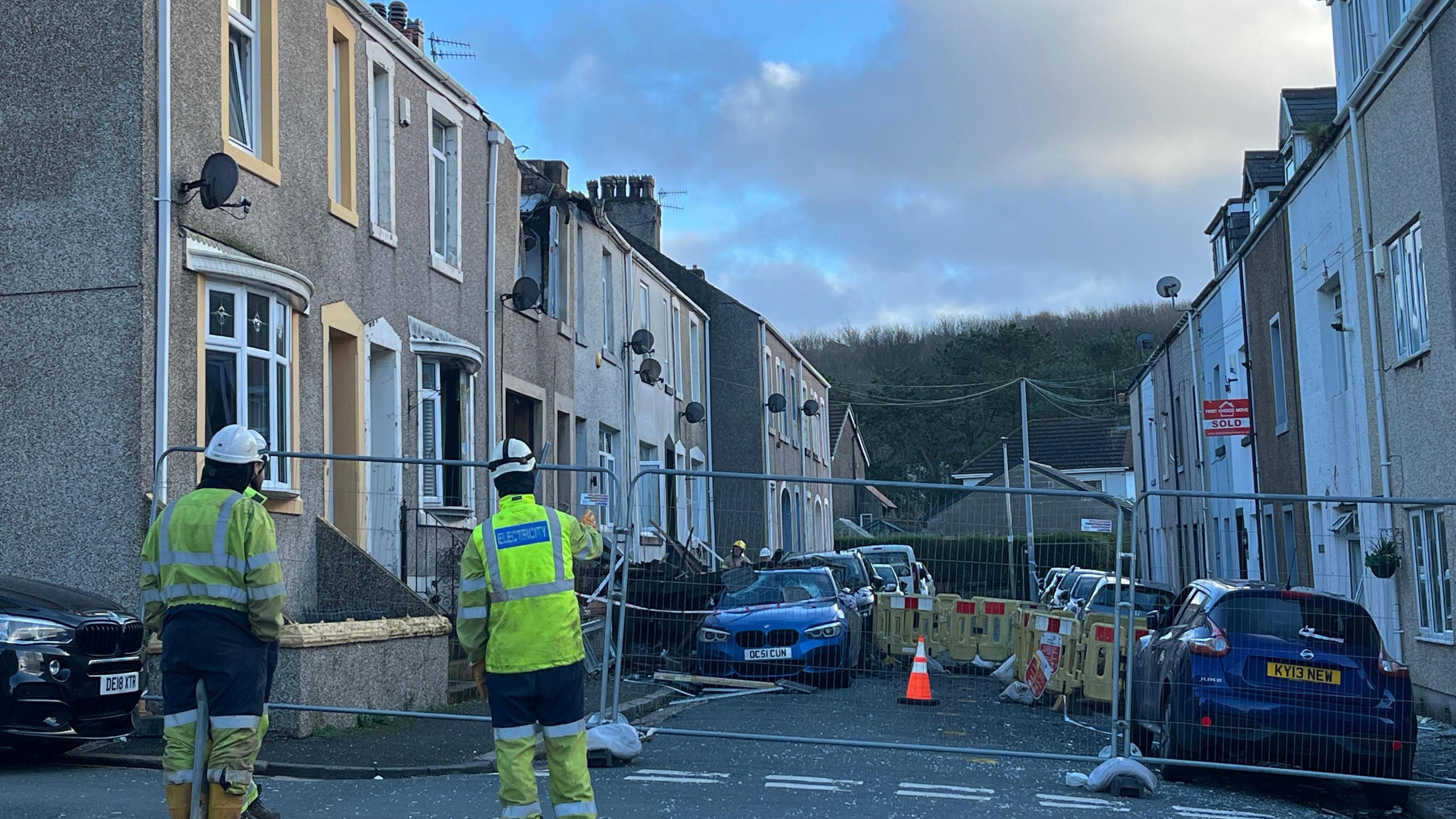 Officers in high-vis jackets stand on a street, which is partly fenced off. A house has visible damage to its first flood and roof and a car parked outside also has its roof and windscreen smashed.