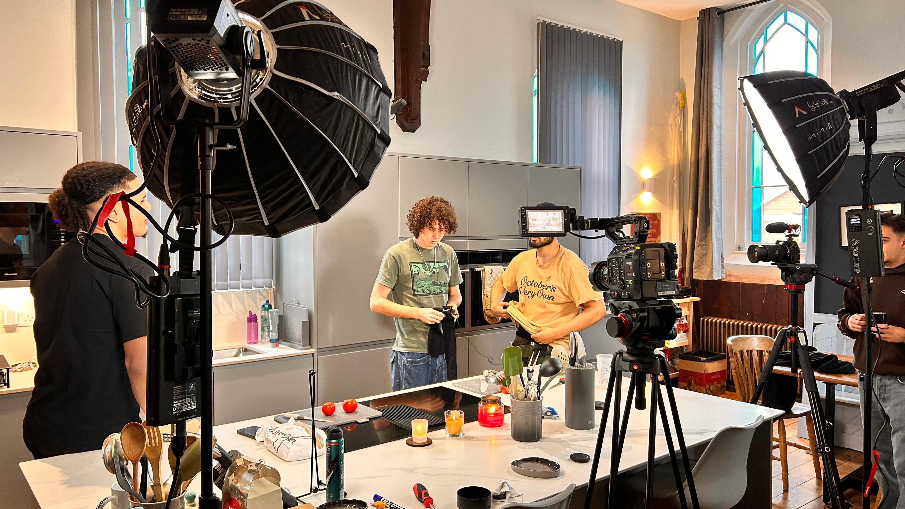 Behind-the-scenes view of a video shoot in a modern kitchen setup, with cameras, lighting equipment, and crew members filming two men at a kitchen island.