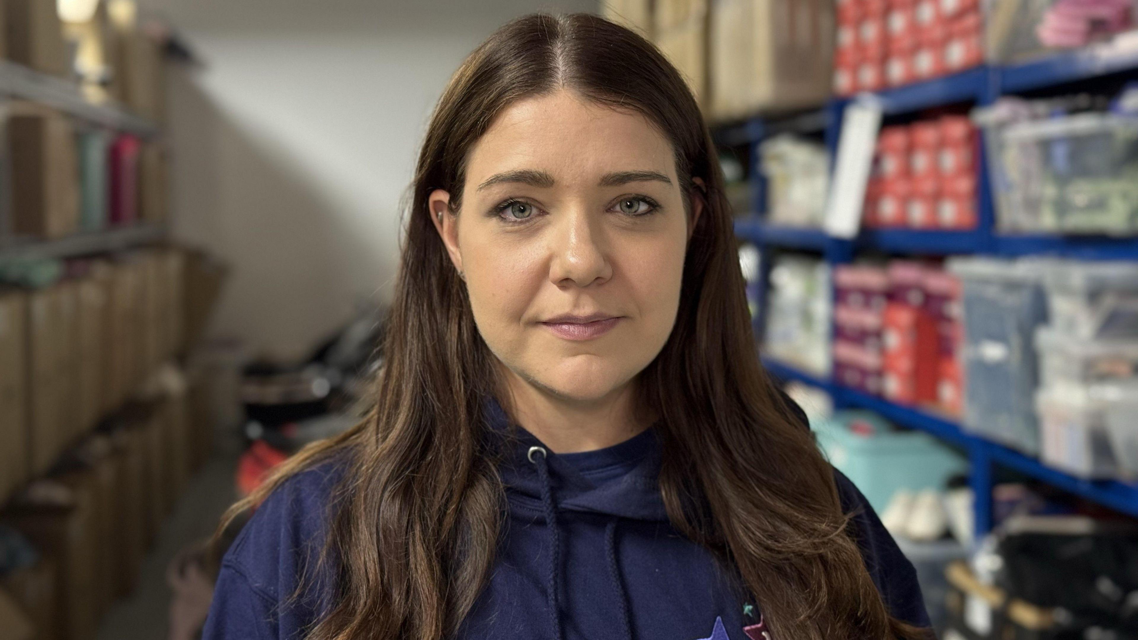A woman with long brown hair is wearing a dark blue hoodie. She is standing in front of shelves that are on the left and right of a narrow room. There are boxes and products stacked on them