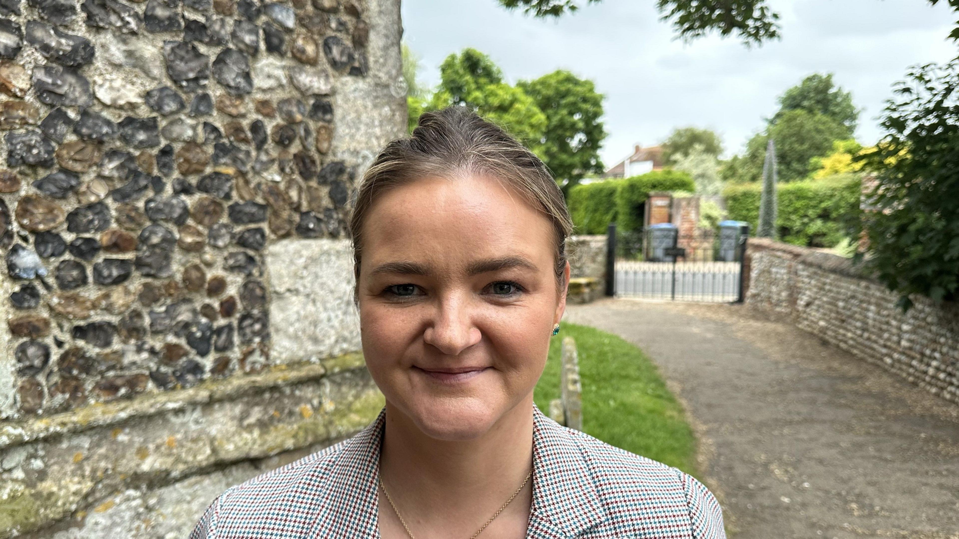 A woman with tied-back hair smiles at the camera. She is wearing a houndstooth-check jacket, white blouse and gold necklace. She is standing in a churchyard with a gate and the chuch wall behind her.