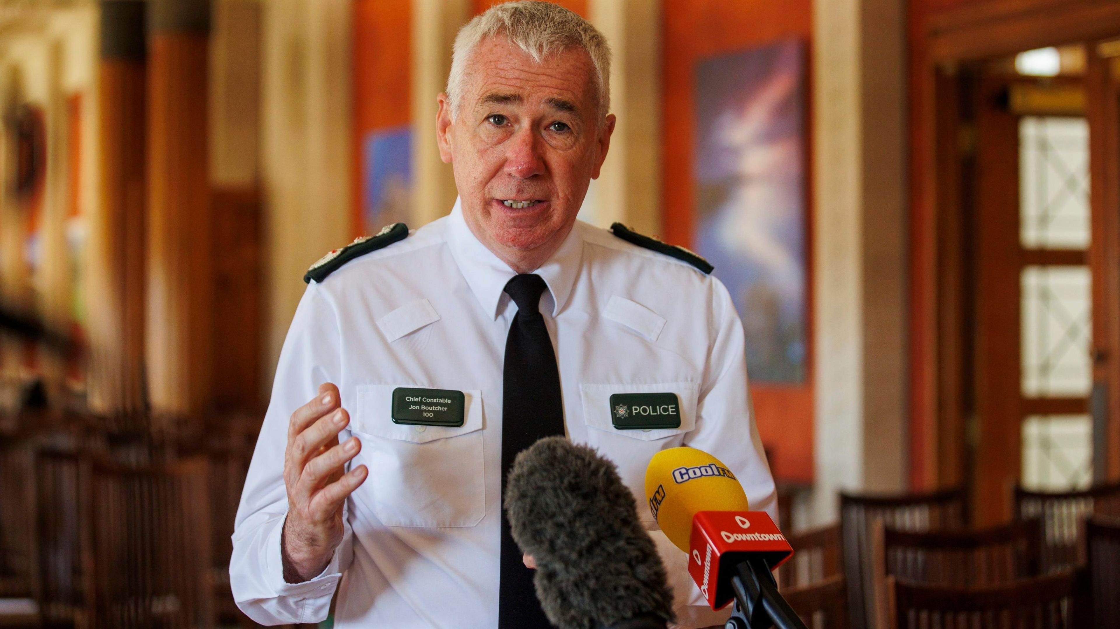 John Boutcher has grey hair and is wearing a white uniform shirt with PSNI insignia on its shoulders and chest. He is gesturing with his right hand and there are two broadcasters' microphones in front of him.