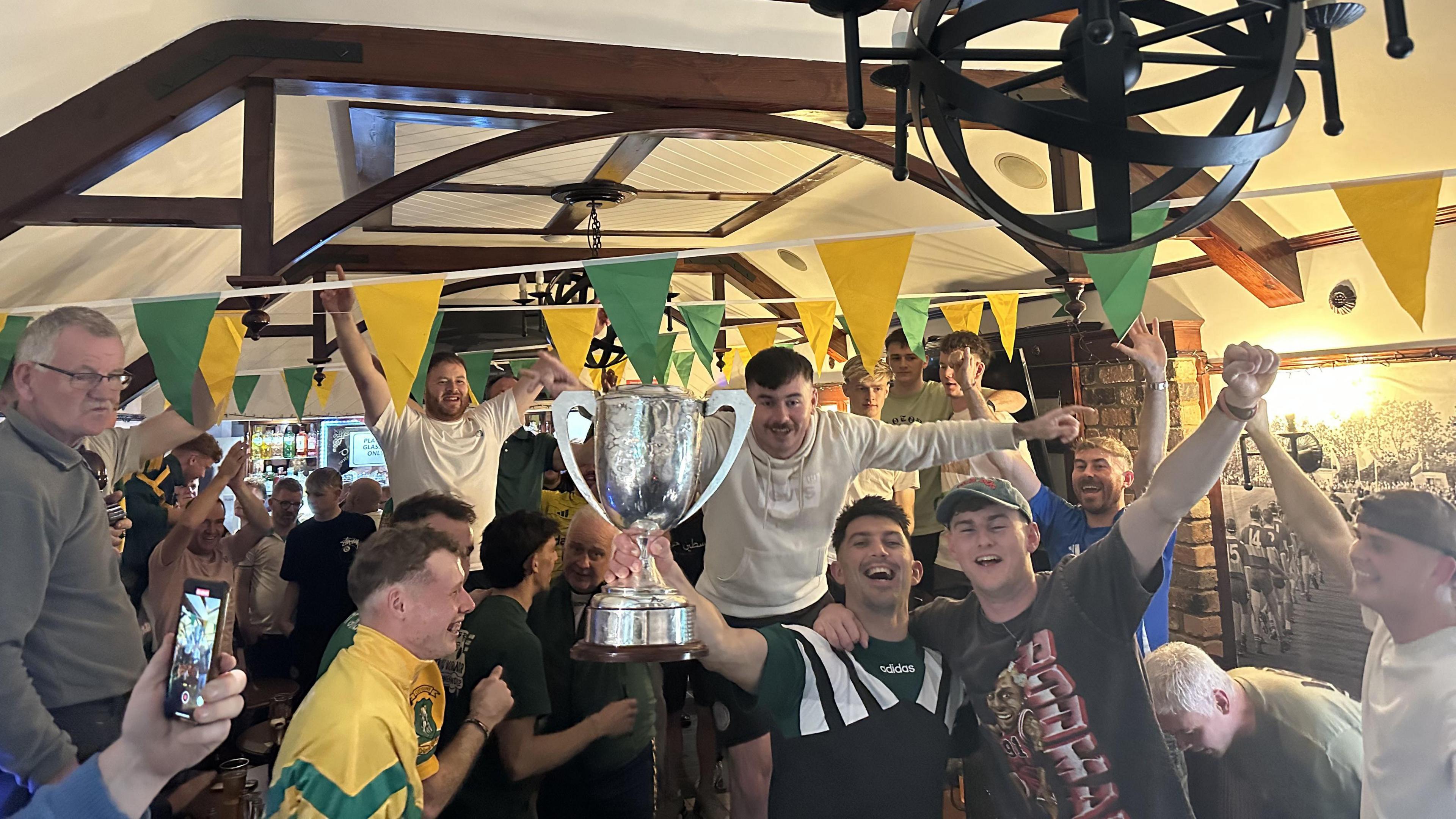 A large group of men celebrating in a pub. One man is holding up a silver trophy. There is yellow and green bunting hanging from the ceiling.