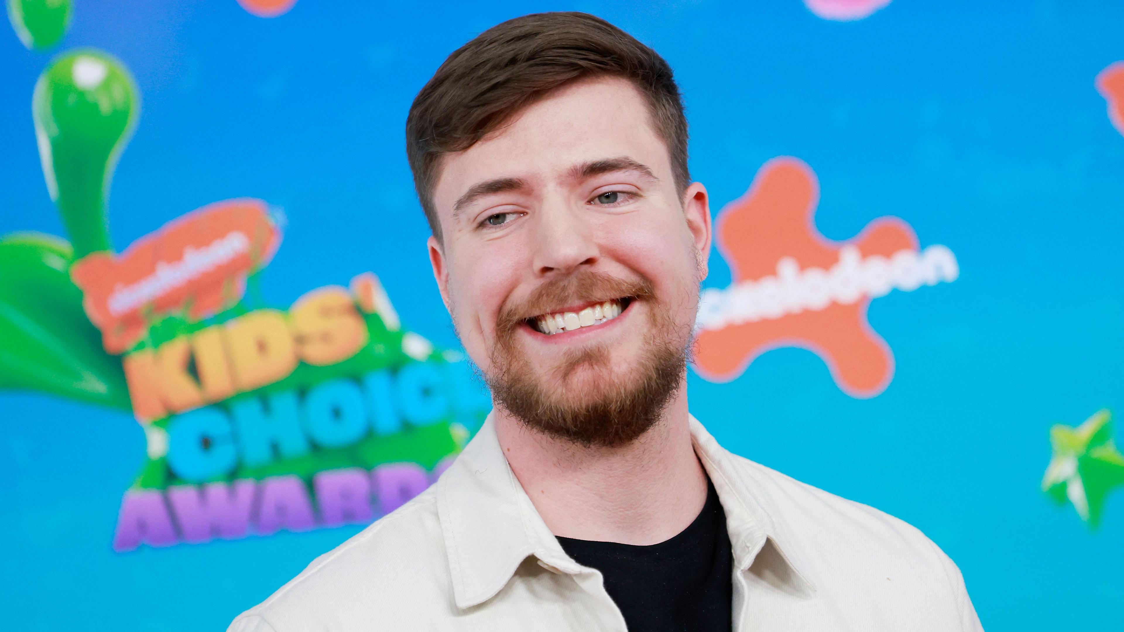 Jimmy Donaldson, aka MrBeast, pictured at the Nickelodeon Kids' Choice Awards. MrBeast has short brown hair and a short beard. He wears a white jacket over a black T-shirt and poses in front of a bright blue hoarding with Nickelodeon's orange branding. 