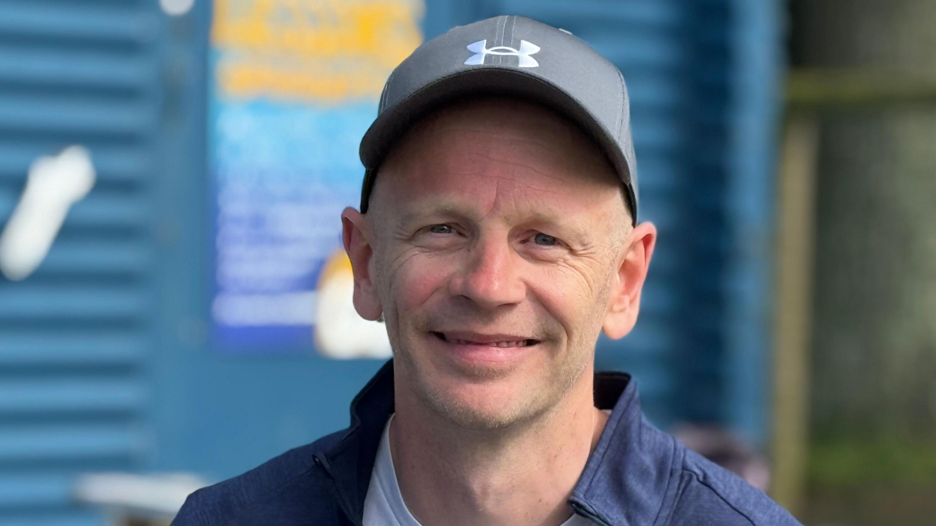 A smiling man in a blue cap in front of a skate park coffee shop.