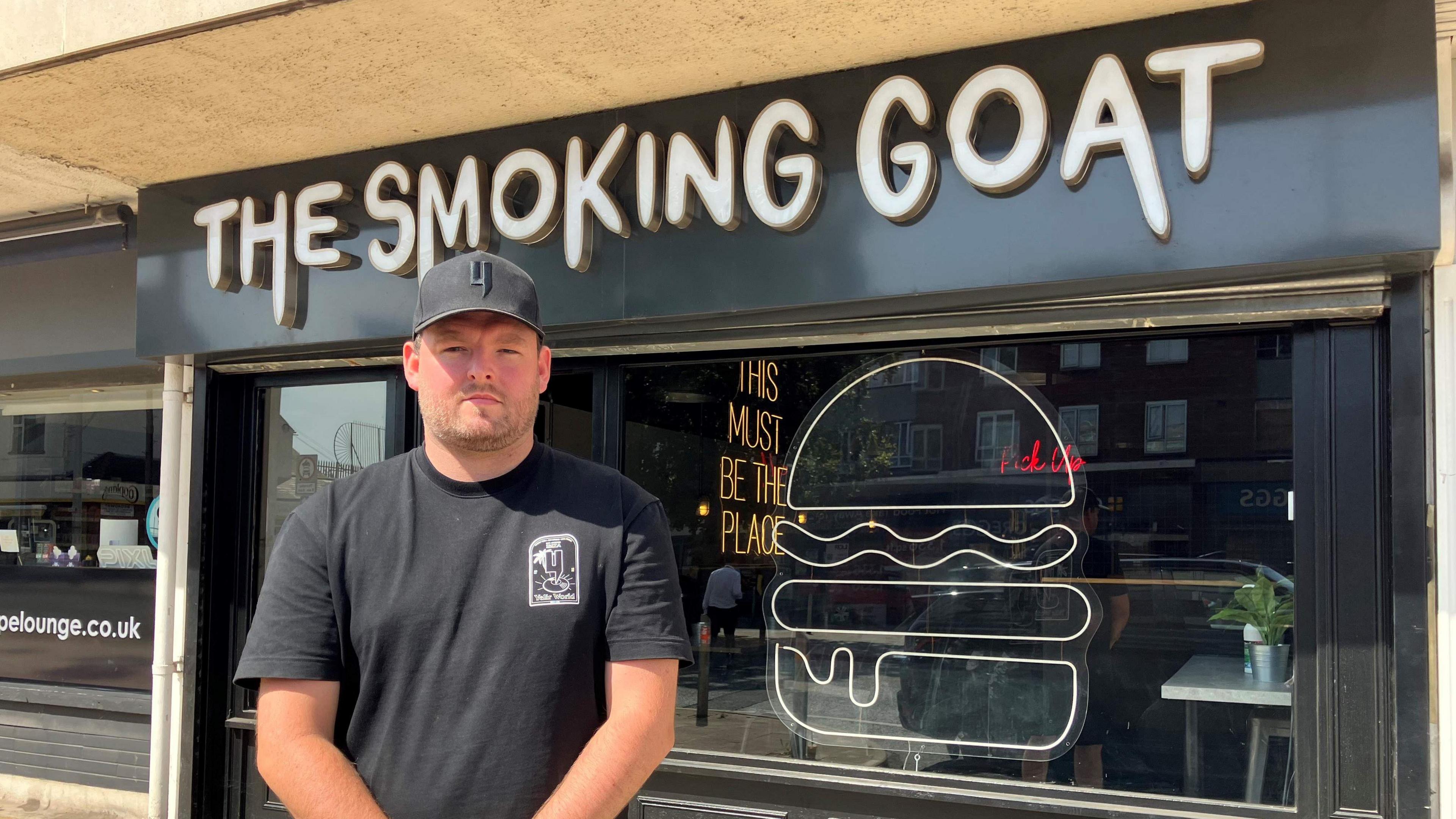 A man wearing a dark cap and a dark T-shirt. He is standing in front of a shop with a sign that reads "The Smoking Goat". There is a sign on the window in the shape of a burger.