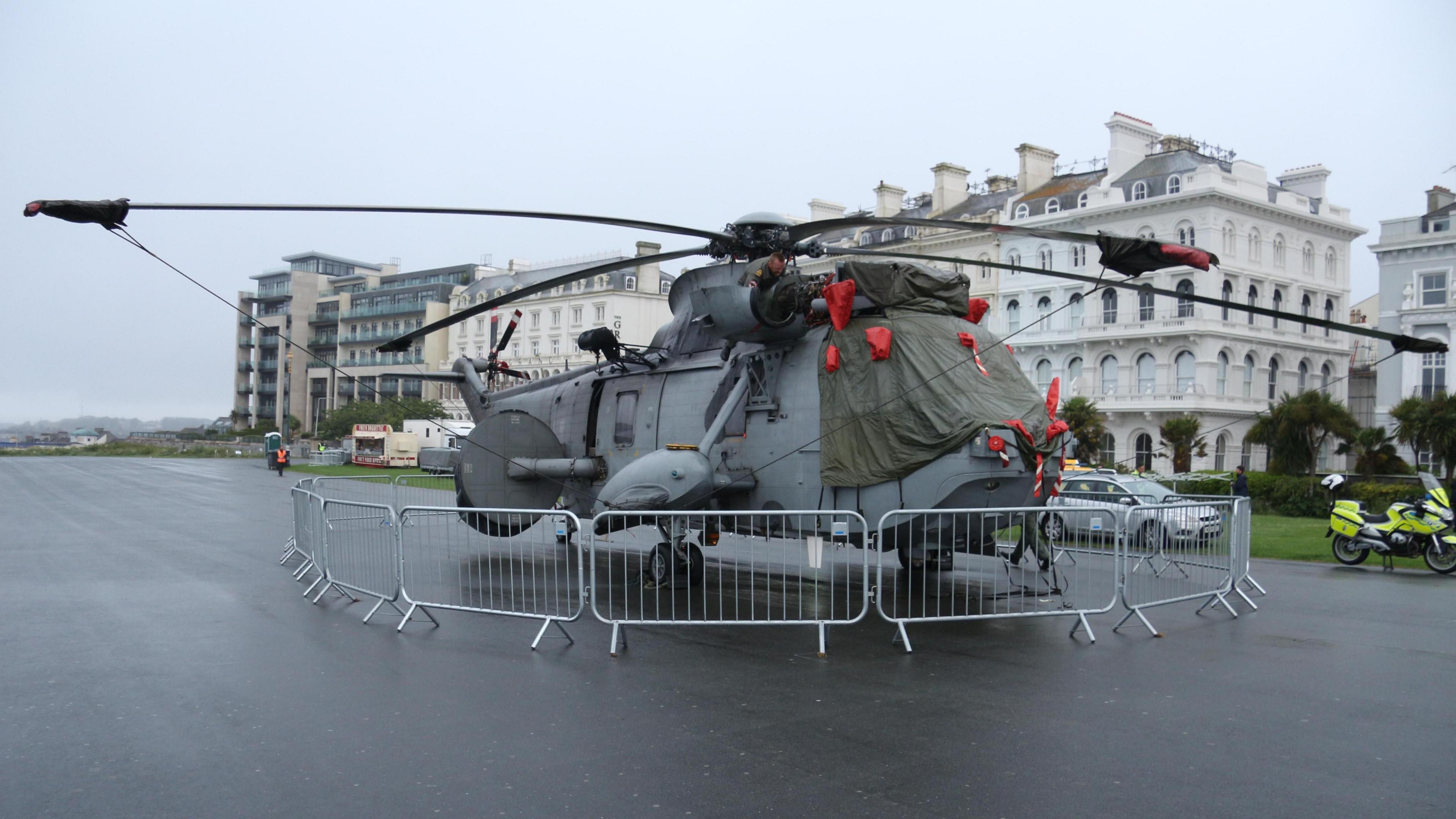 A grey military helicopter on Plymouth Hoe. A police motorbike is to the side of the helicopter. It is a gloomy day.
