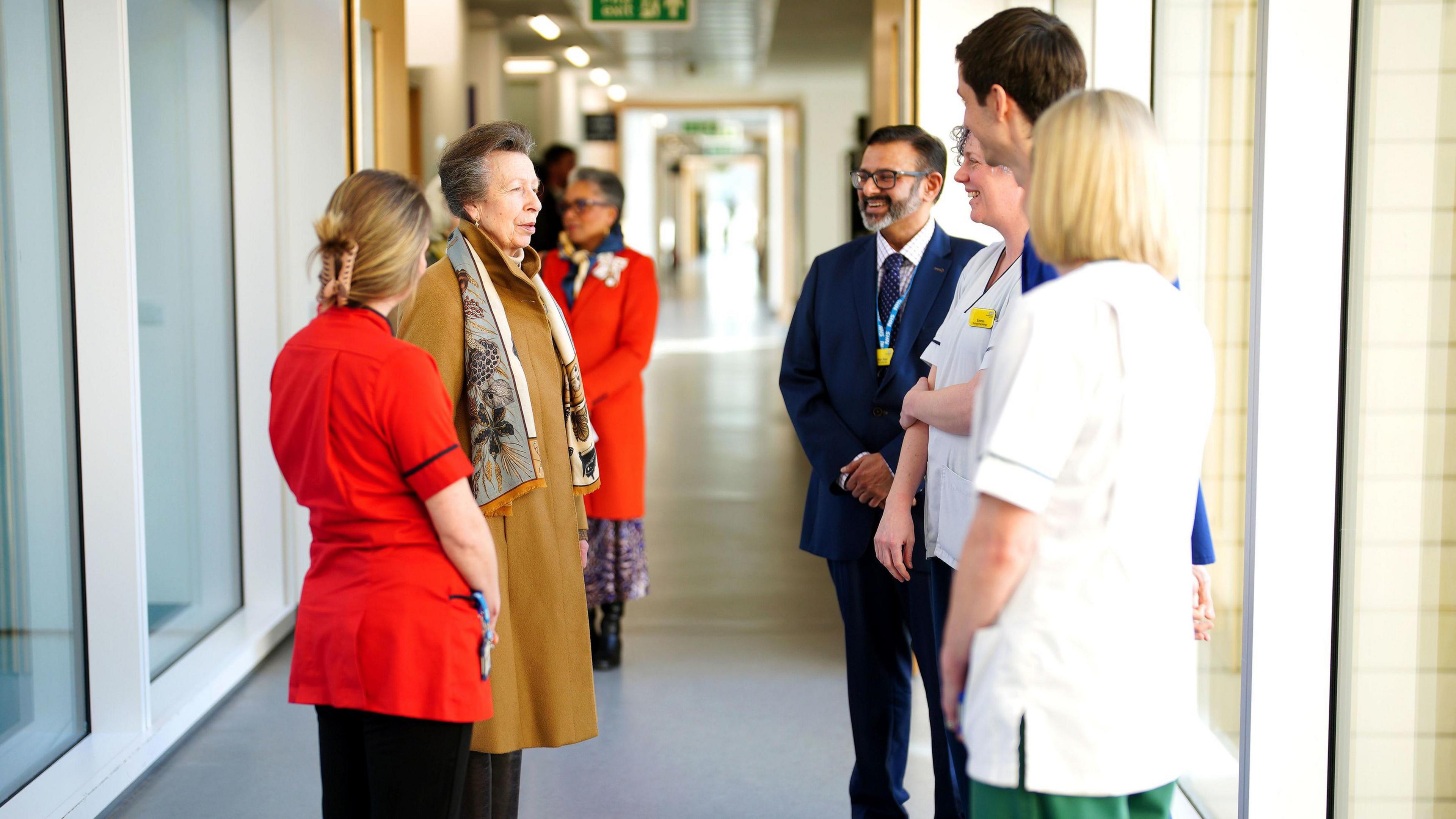 Princess Anne meets clinicians during her visit. There are five people standing around her in a semi-circle, and they are all in a light hallway. She is wearing a brown coat and a patterned scarf, while the people around her are either in suits or clinical uniform. 

