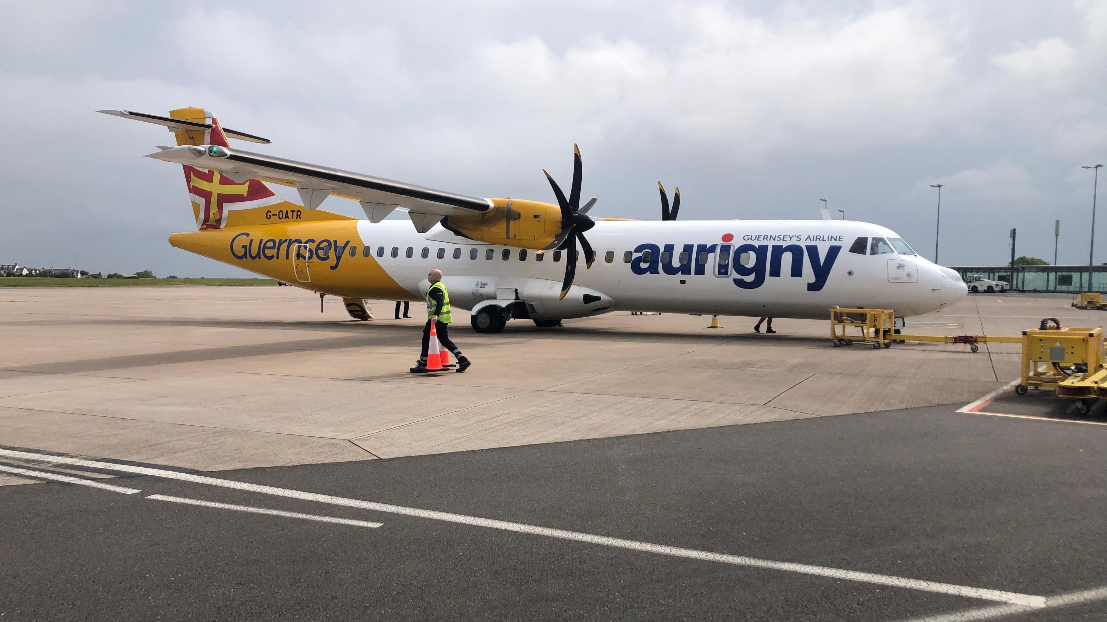 A white propellor plane with a yellow tail and a Guernsey flag. The word Aurigny is written on the side. A man is walking past the wing in a yellow high-vis jacket with two traffic cones, one in either hand. 