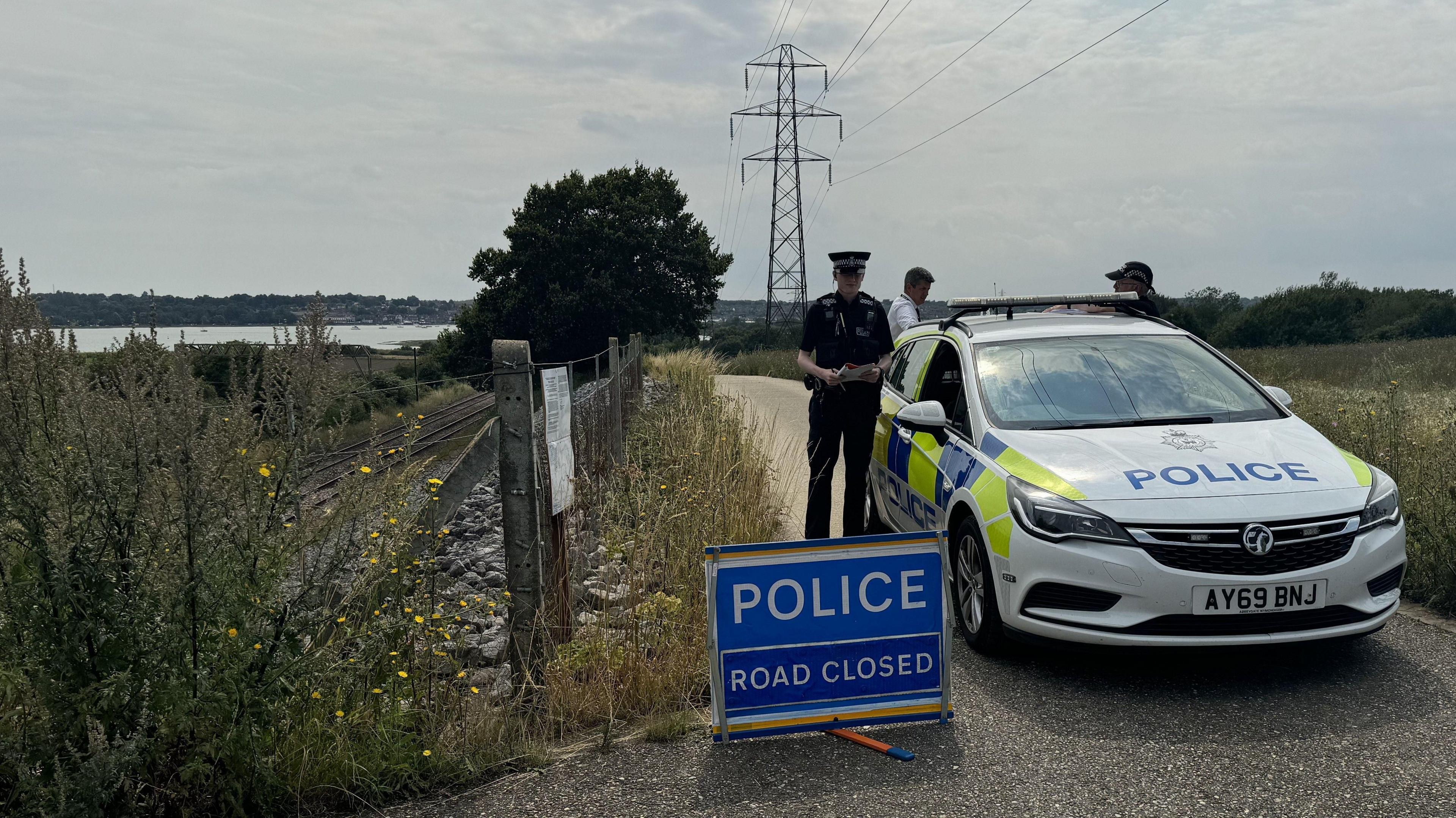 A police car is parked on a metalled track with a sign in front of it saying "POLICE" and "ROAD CLOSED". A police officer is next to the car, with two other people standing behind it. A pylon is in the field further along the track. The railway line and River Stour estuary are on the left of the picture.