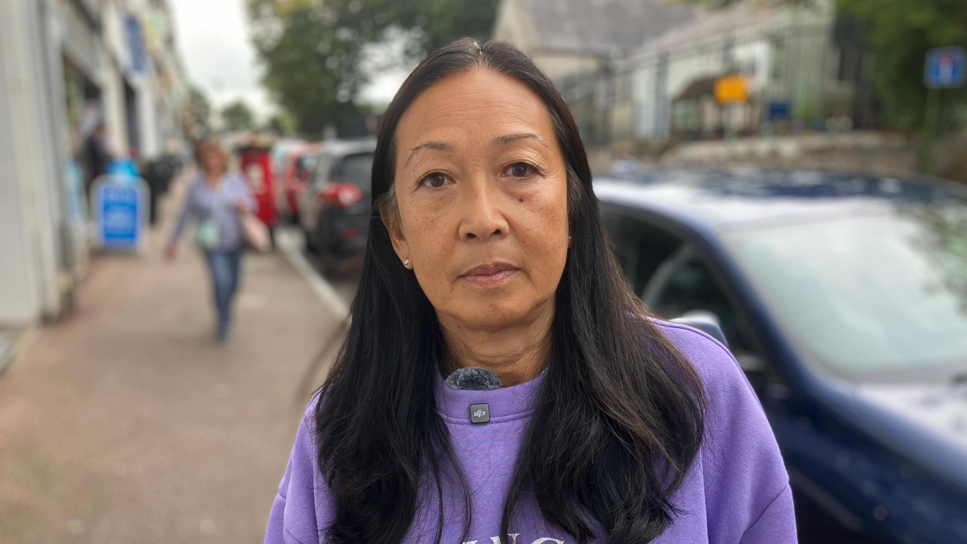 A woman in a purple sweatshirt stands on a pavement alongside a row of shops on her right with parked cards on her left