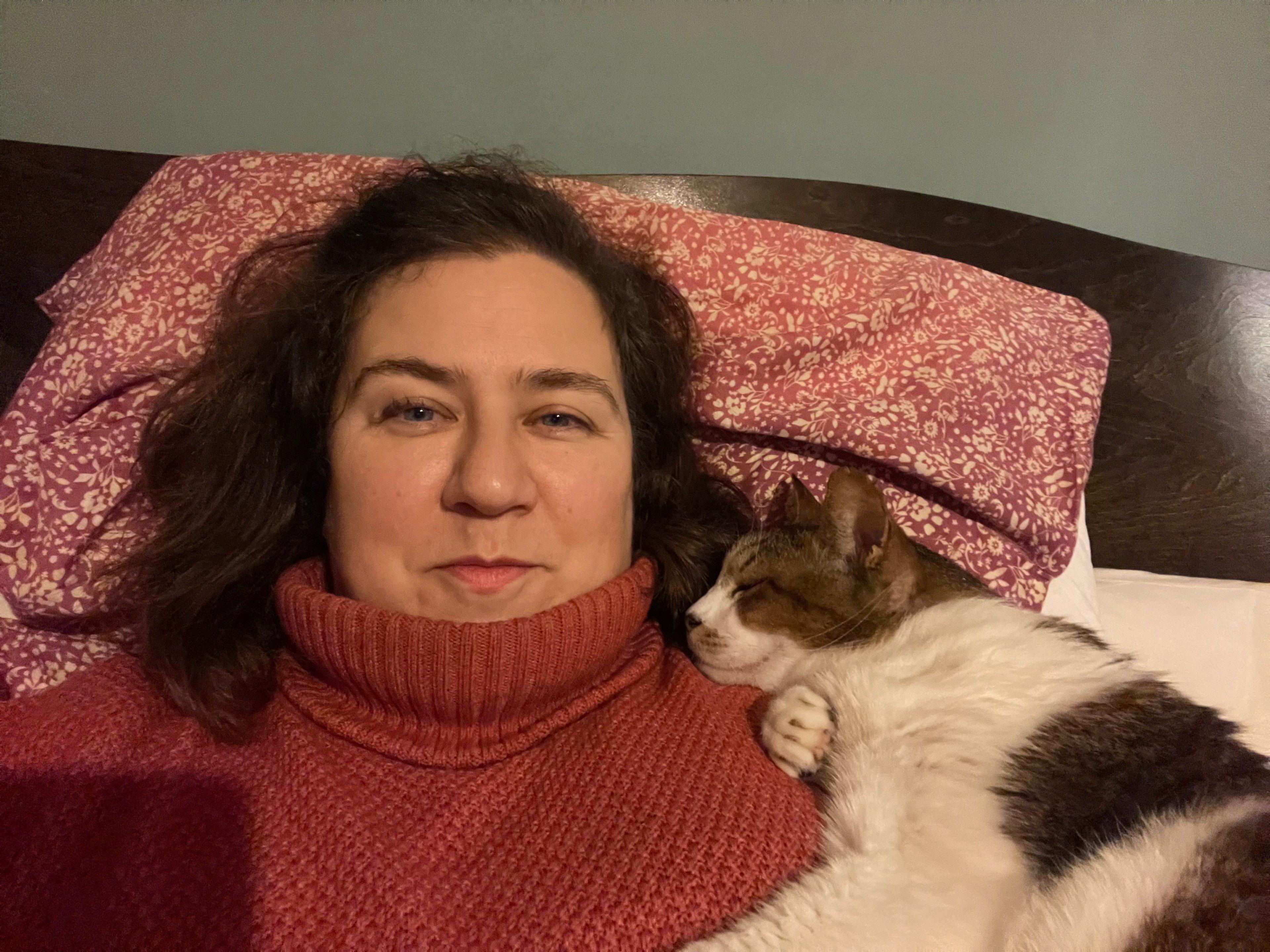 A woman wearing a red jumper lays on a bed, her head on a pillow. A brown and white cat nuzzles into her shoulder.