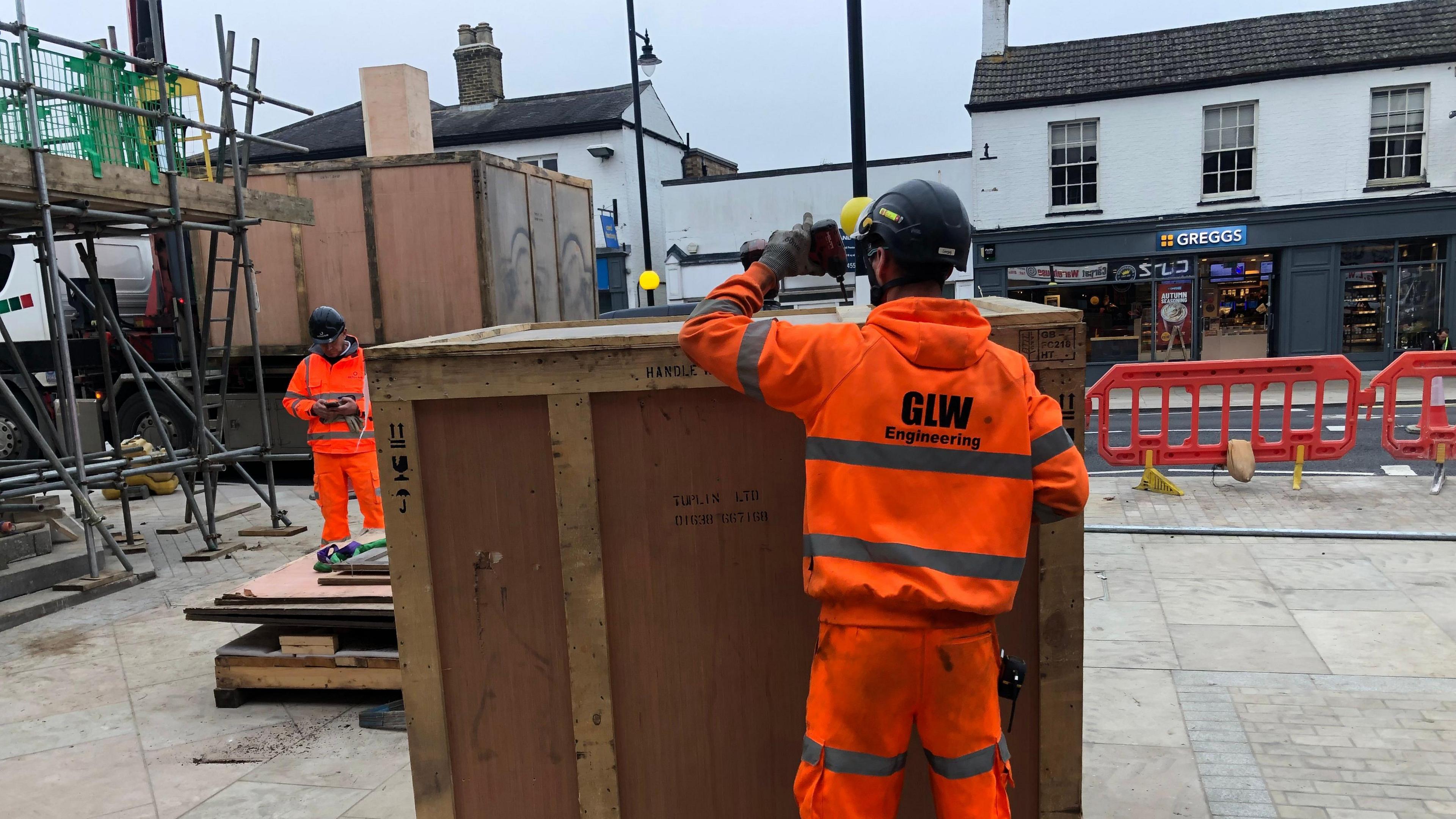 A workman in an orange high-vis jacket and trousers uses a cordless drill to remove screws on one of the crates containing parts of the Fountain, a much larger box can be seen on the back of a flat bed lorry.