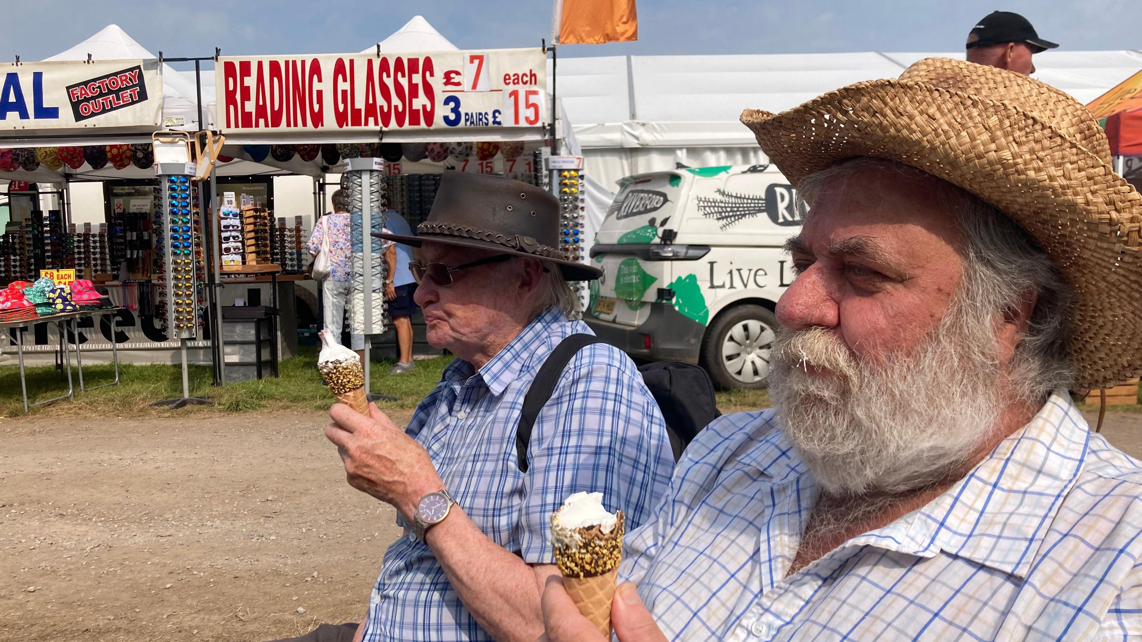 Two men in hats and checked short sleeve shirts enjoying ice creams with a stall selling glasses and a Riverford-branded van in the background