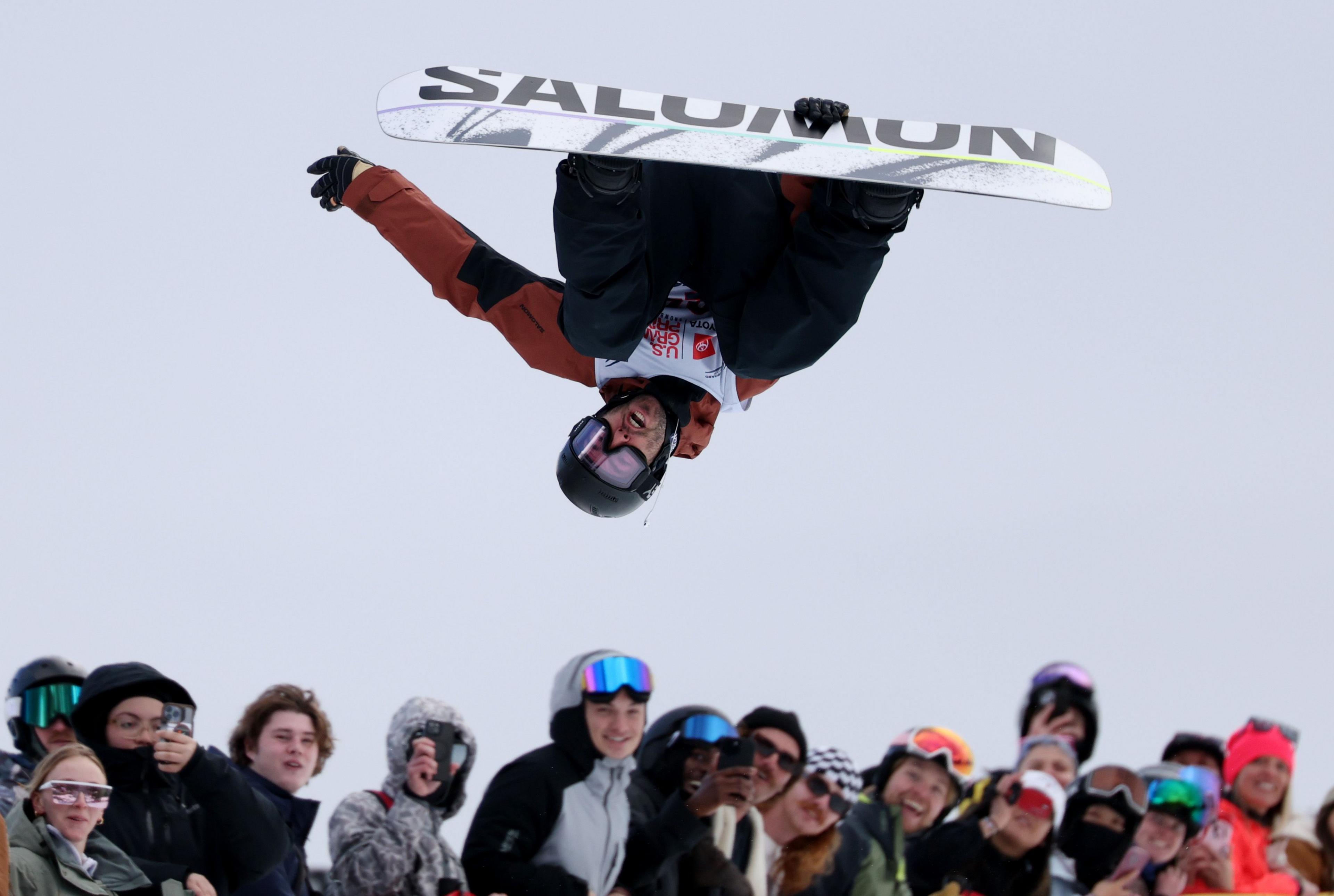 A snowboarder performs an inverted aerial trick high above a crowd of spectators, holding a white board with bold lettering while spinning upside down. The audience below, bundled in winter clothing and helmets, watches the jump against an overcast sky.