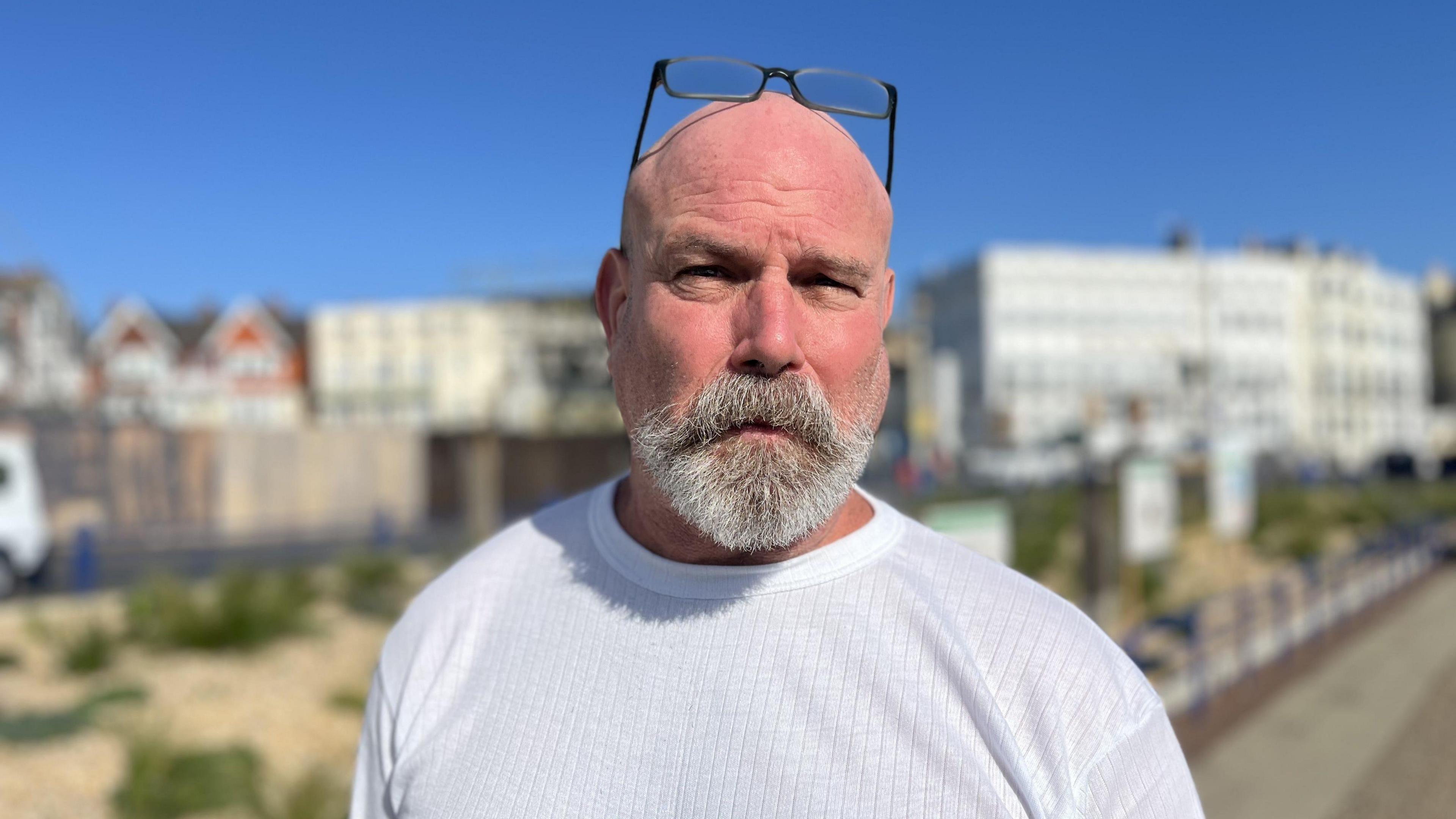 A man in a light grey t-shirt with his glasses on his head. In the background is a row of hoardings around the former site of the Claremont Hotel.